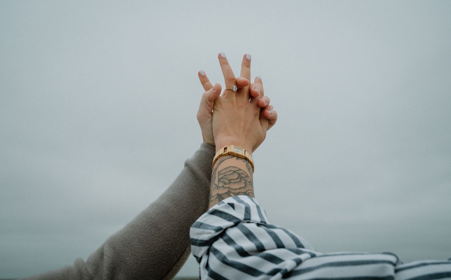 Two hands intertwined against a cloudy sky, one adorned with a gold watch and a tattoo.