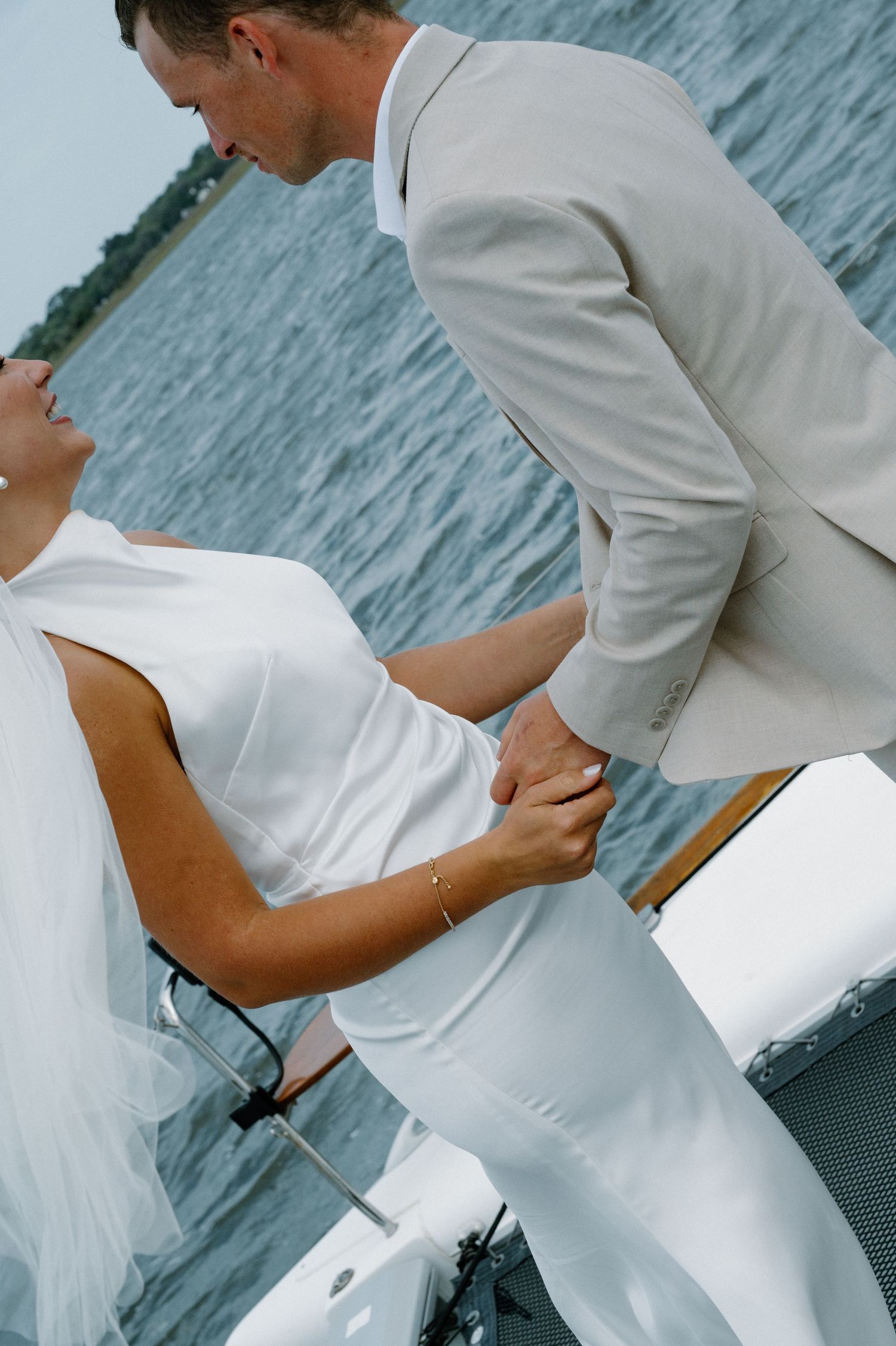 Bride and groom holding hands on a boat near the water, wearing white wedding dress and beige suit.