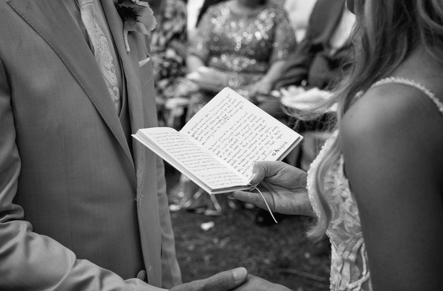 Close-up black and white photo of groom reading handwritten wedding vows from a small notebook.