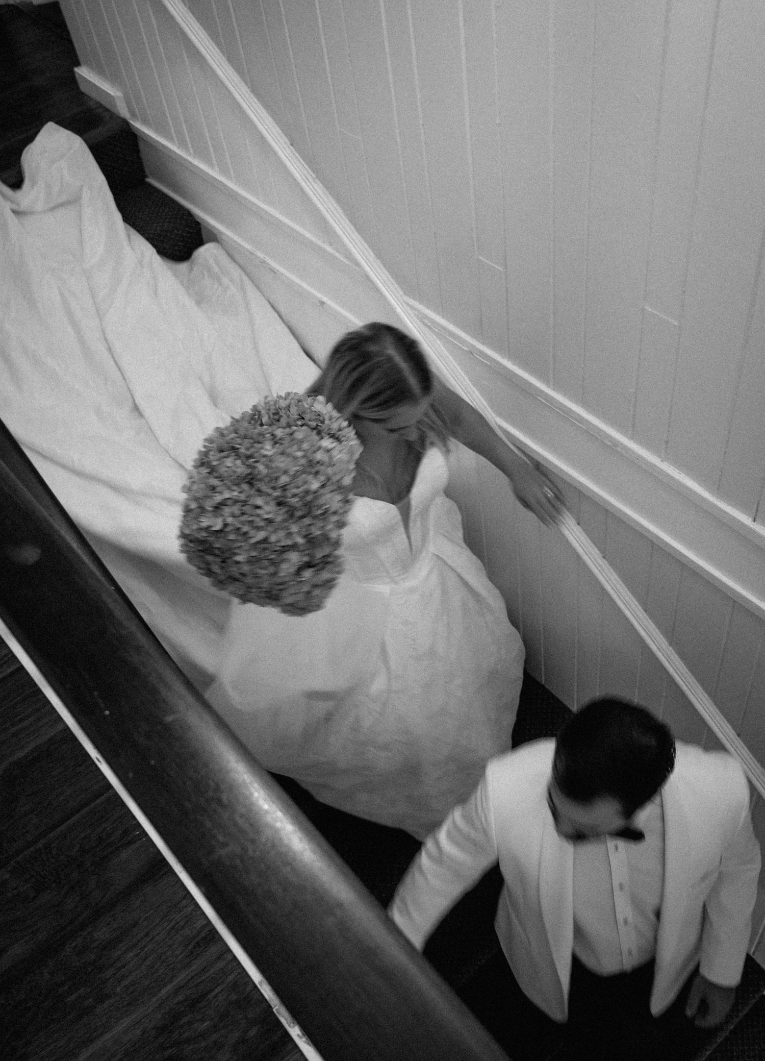 Aerial black and white view of bride descending narrow staircase with flowing gown and bouquet.
