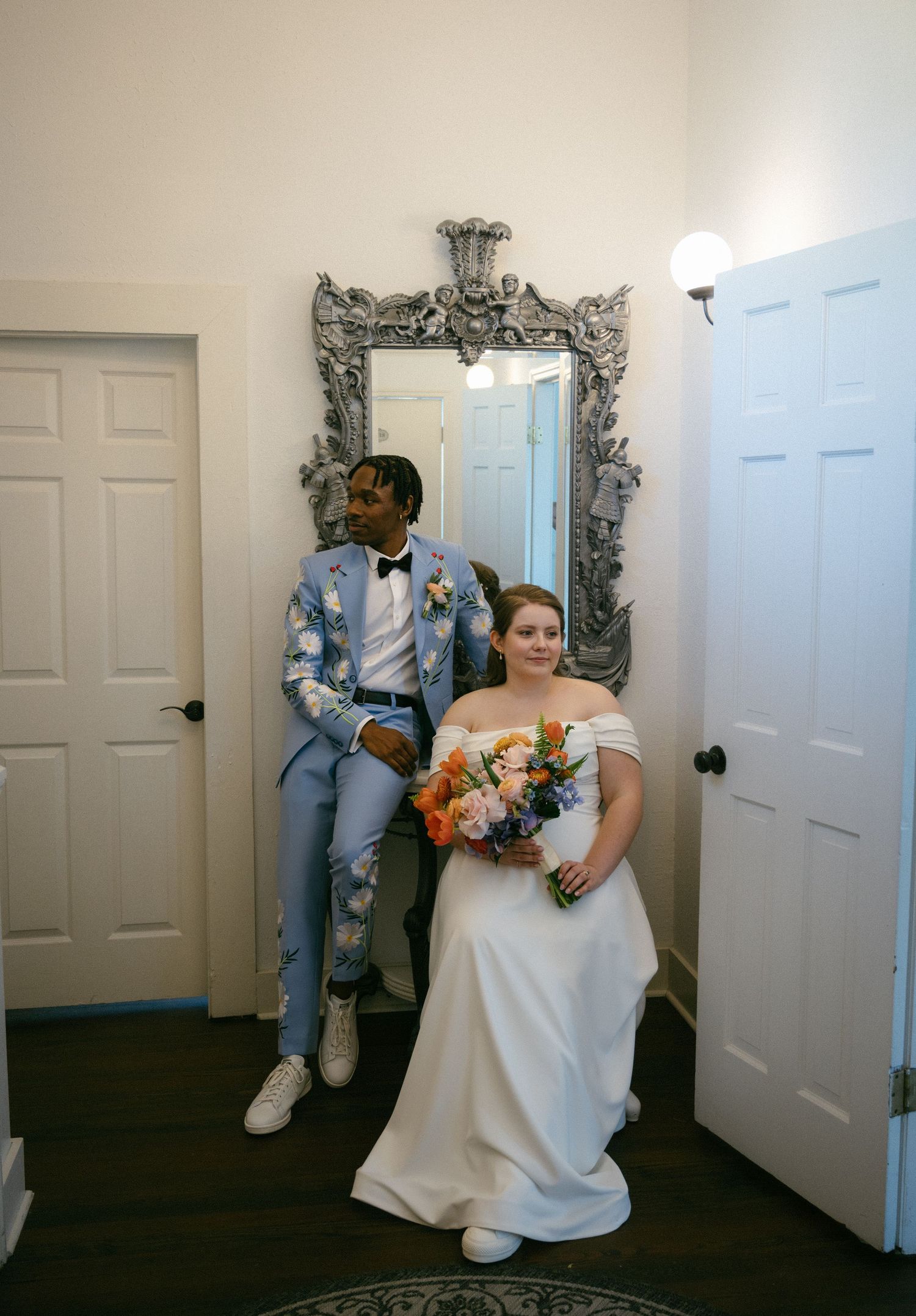 Bride holding colorful bouquet and groom in light blue suit pose before ornate silver mirror in white room.