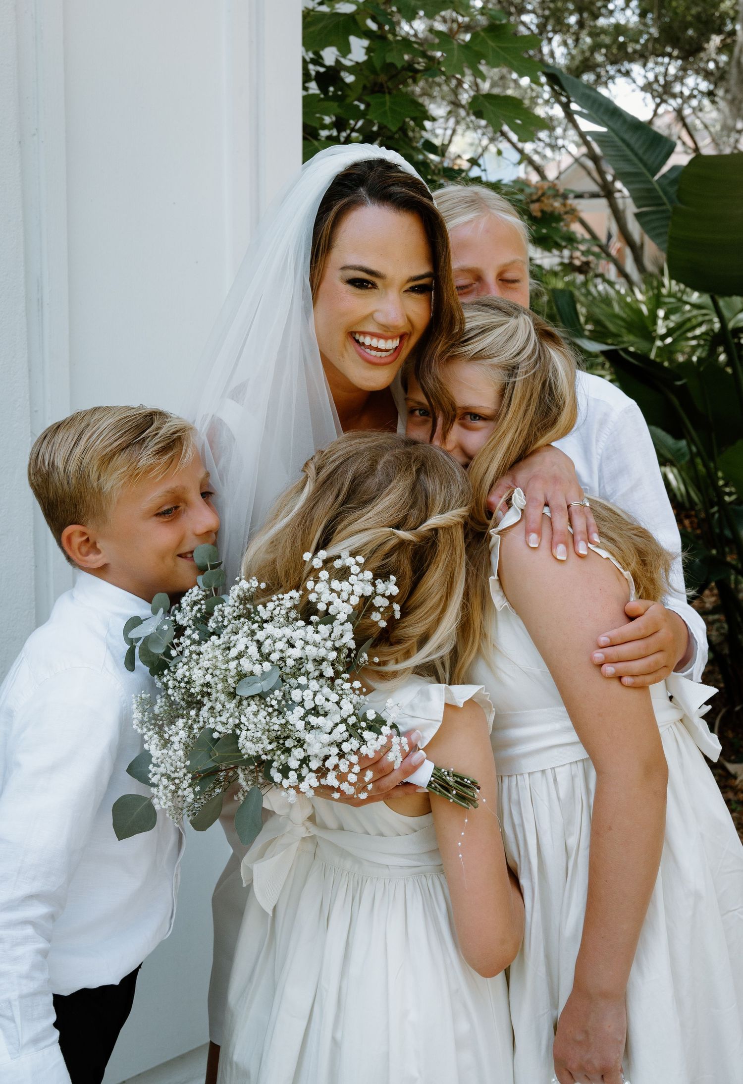 Smiling bride in white veil embraced by children in white outfits holding baby's breath bouquet outdoors.