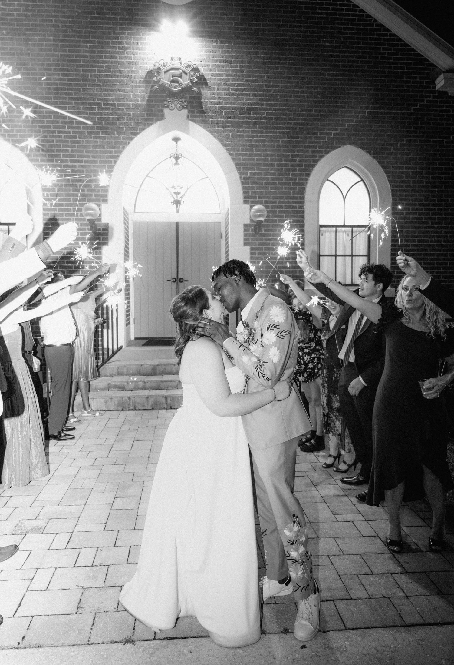 Black and white photo of newlyweds kissing during sparkler send-off outside brick chapel at night in Jacksonville, Florida.