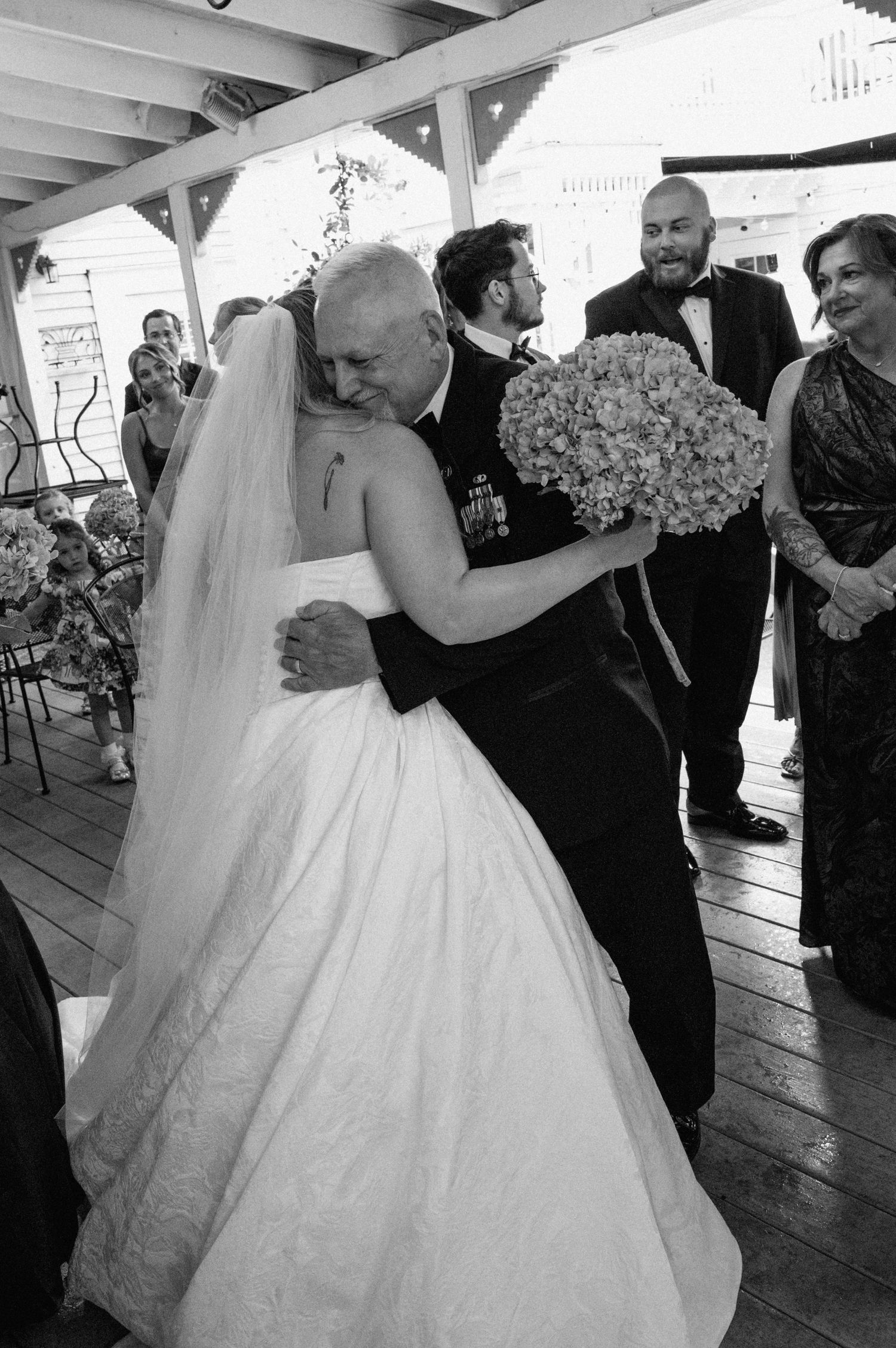 Emotional black and white photo of bride hugging father during wedding ceremony on wooden deck.