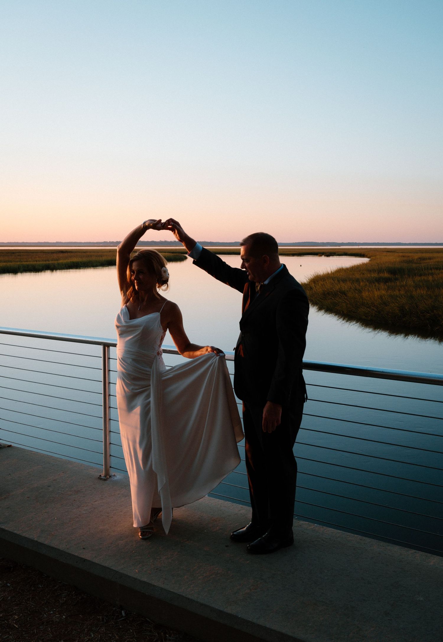Bride and groom forming a heart shape with their hands at sunset over a calm waterway.
