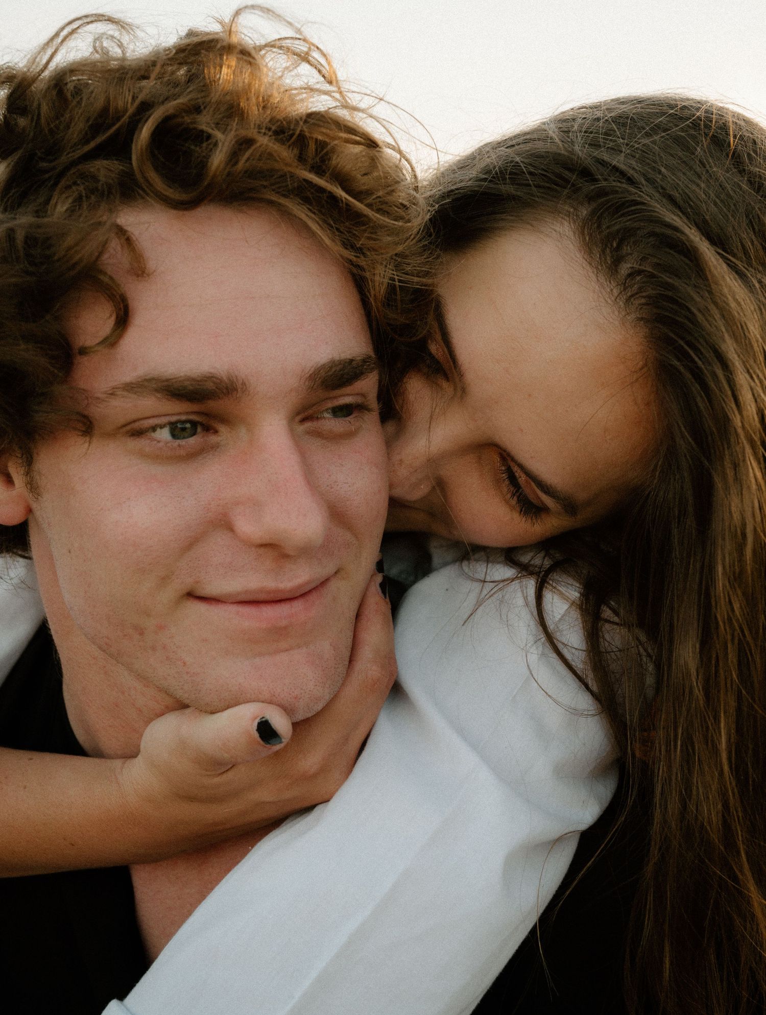 Young couple sharing a tender moment, woman embracing man from behind and kissing his cheek outdoors.