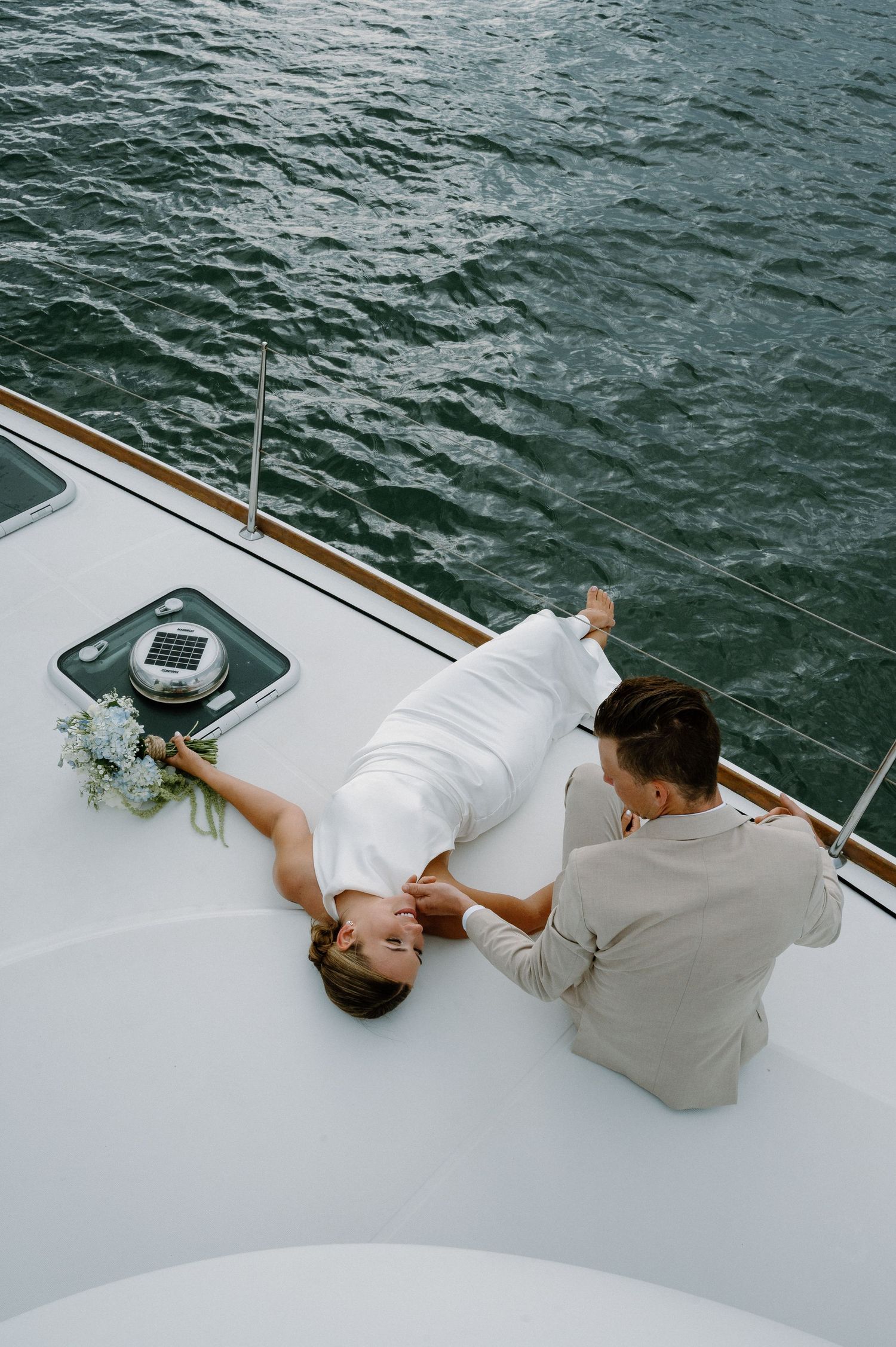 Bride and groom lying on the deck of a white catamaran with ocean water visible below.