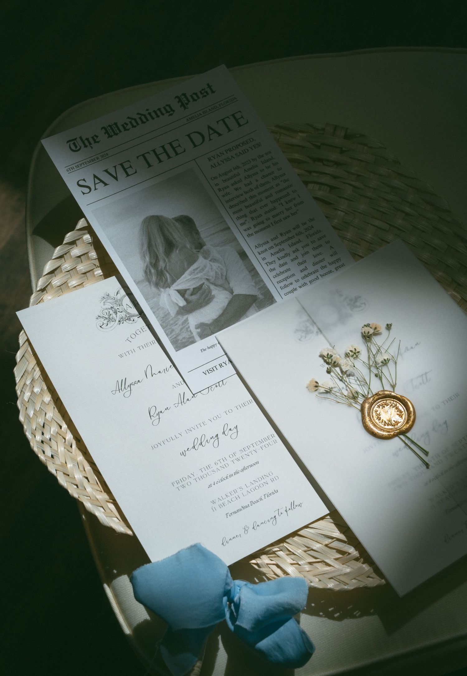 Elegant wedding stationery including a newspaper-style save the date, invitation, and wax seal on a wicker tray.