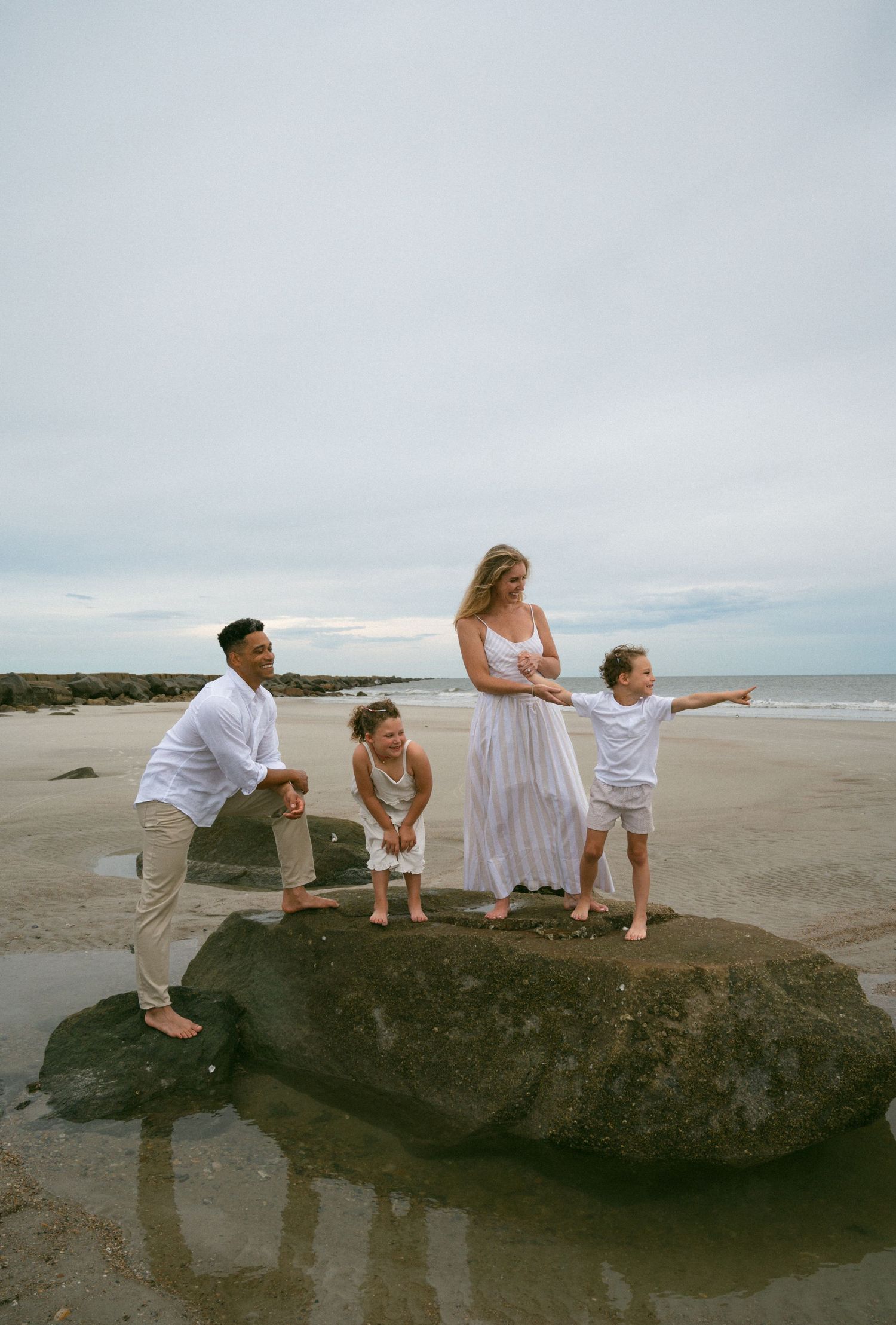Family of four posing on large rock on sandy beach, children pointing toward the ocean.