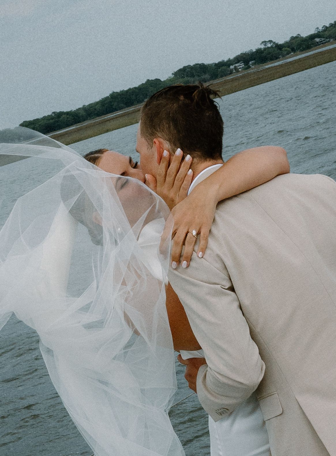 Bride and groom sharing a romantic kiss by the water, her veil flowing in the wind.