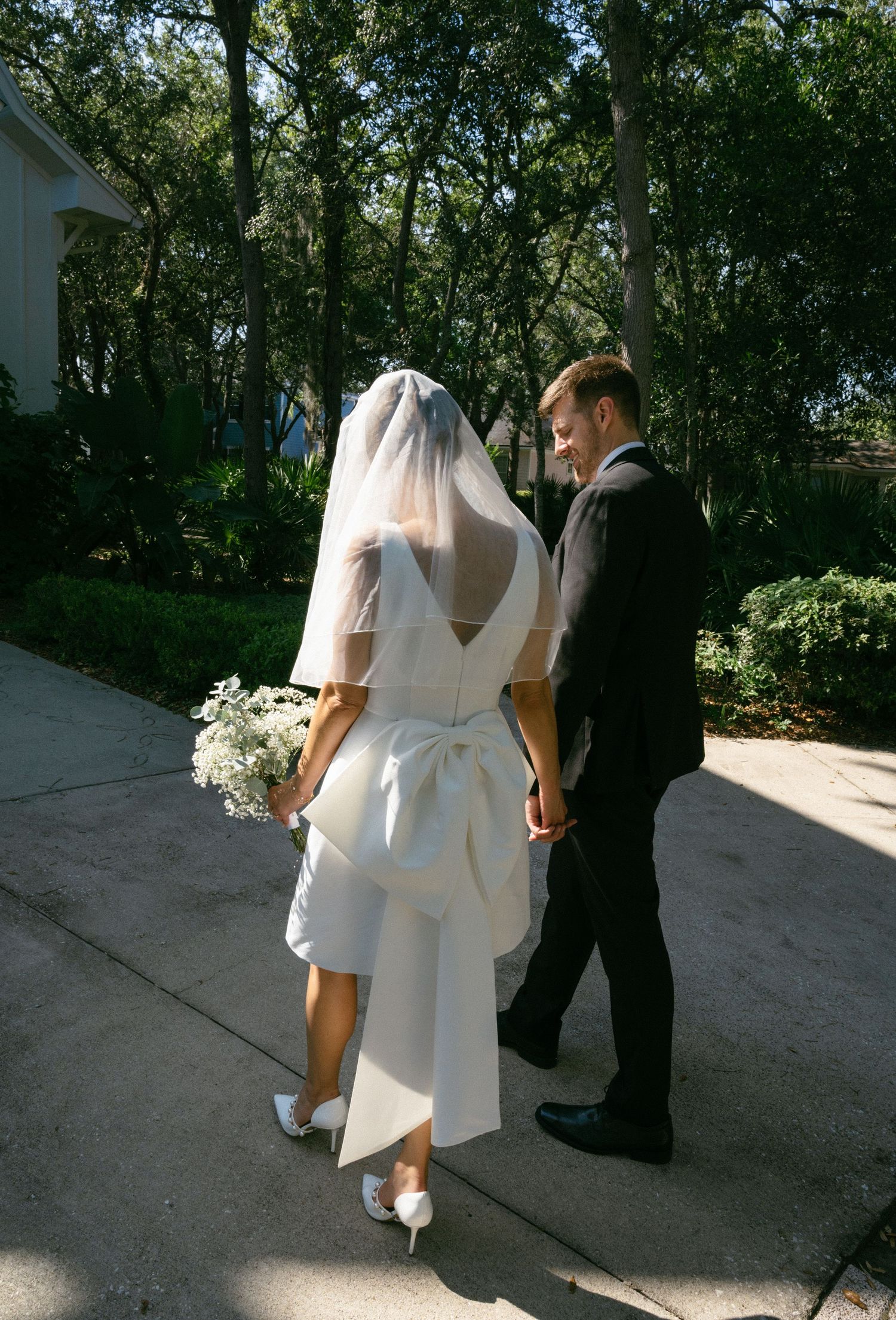Bride and groom walking hand in hand on a sunny path, bride wearing white gown with bow and veil holding bouquet.