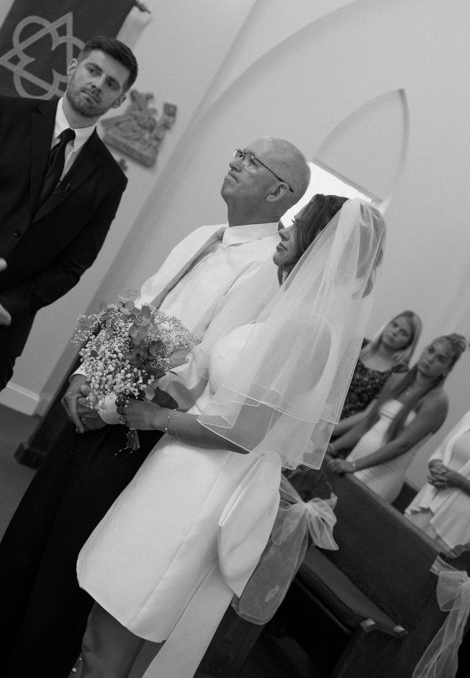 Black and white photo of bride in veil holding bouquet walking down church aisle with escort.