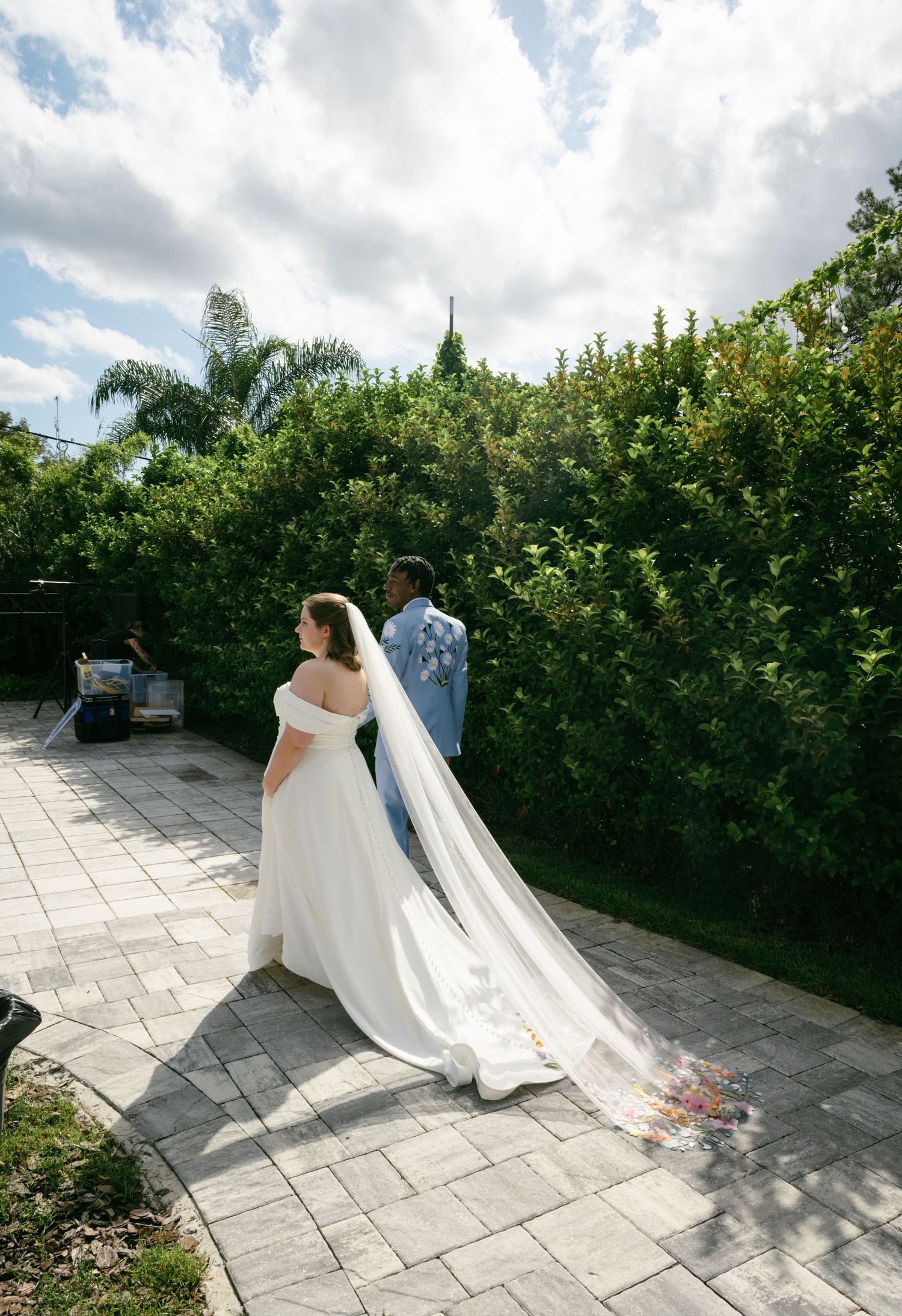Bride in off-shoulder gown with floral cathedral veil walks with groom in blue suit in garden.