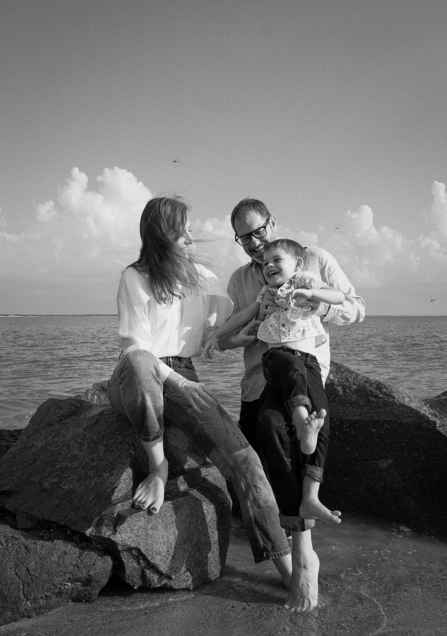 Black and white photo of family with child sitting on rocks by the ocean shore.