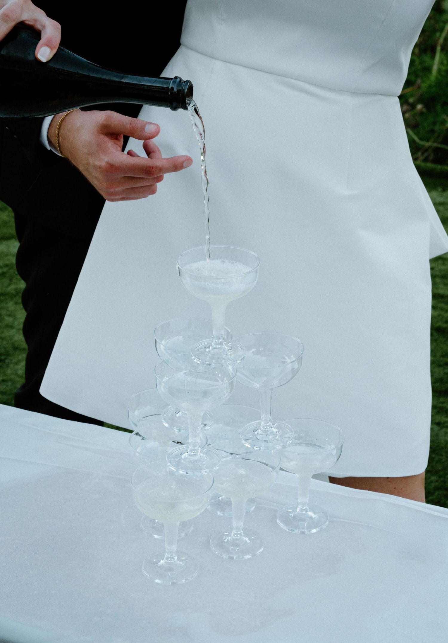 Champagne being poured into a pyramid tower of coupe glasses at an outdoor wedding reception.