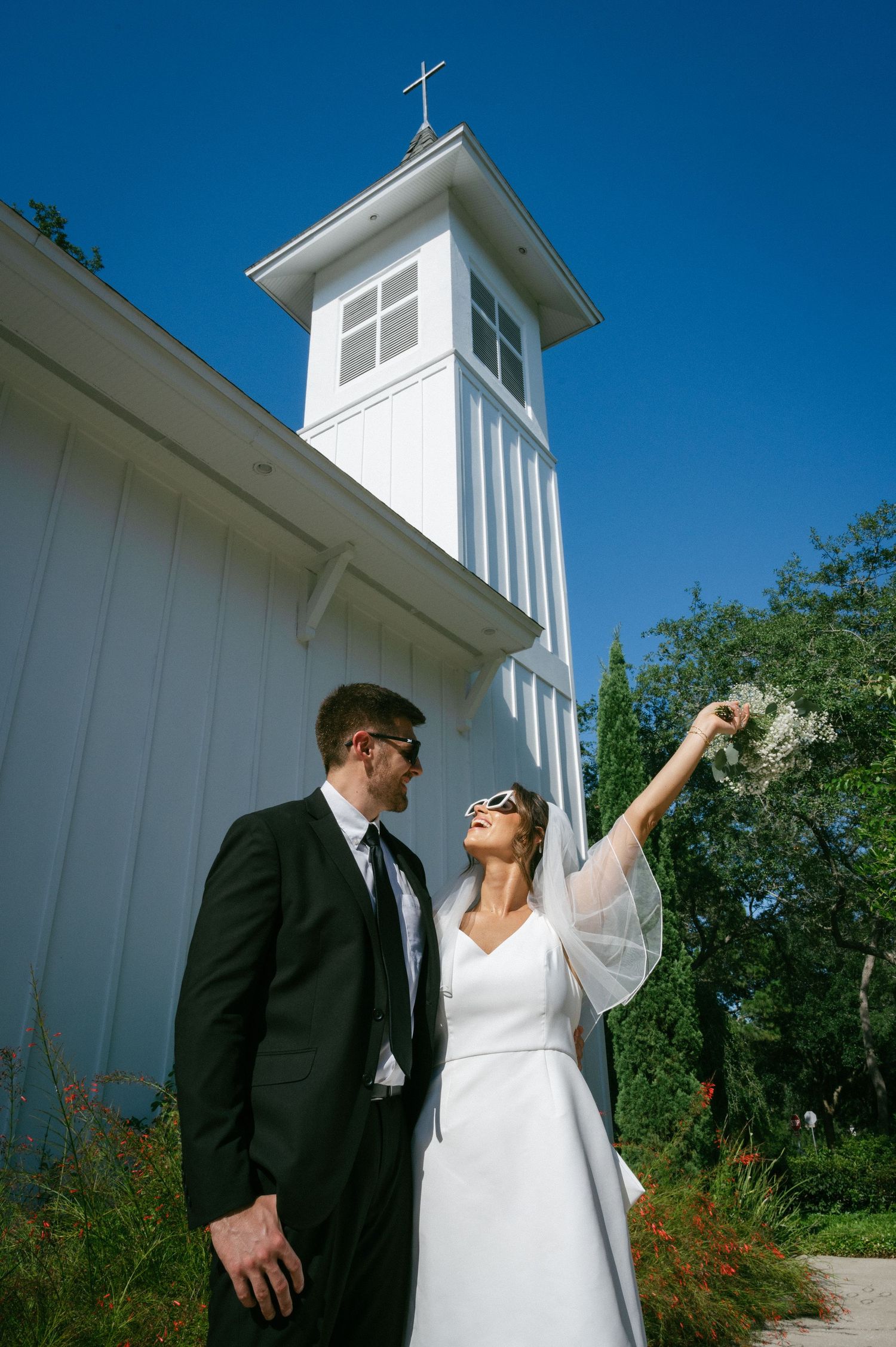 Bride raising bouquet while groom looks on beside white chapel tower against bright blue sky.