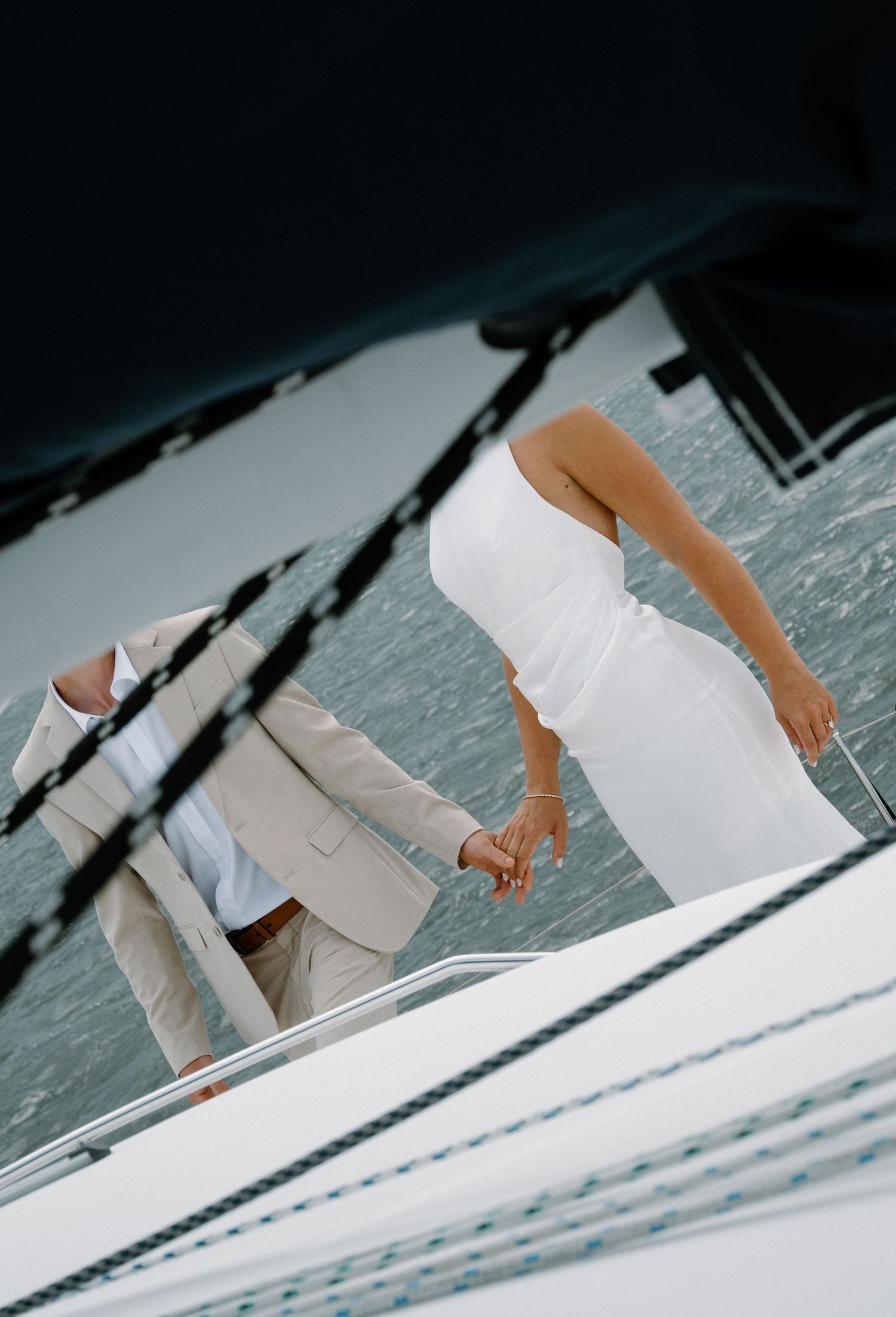 Couple in white dress and beige suit holding hands on a yacht deck with ocean in background.