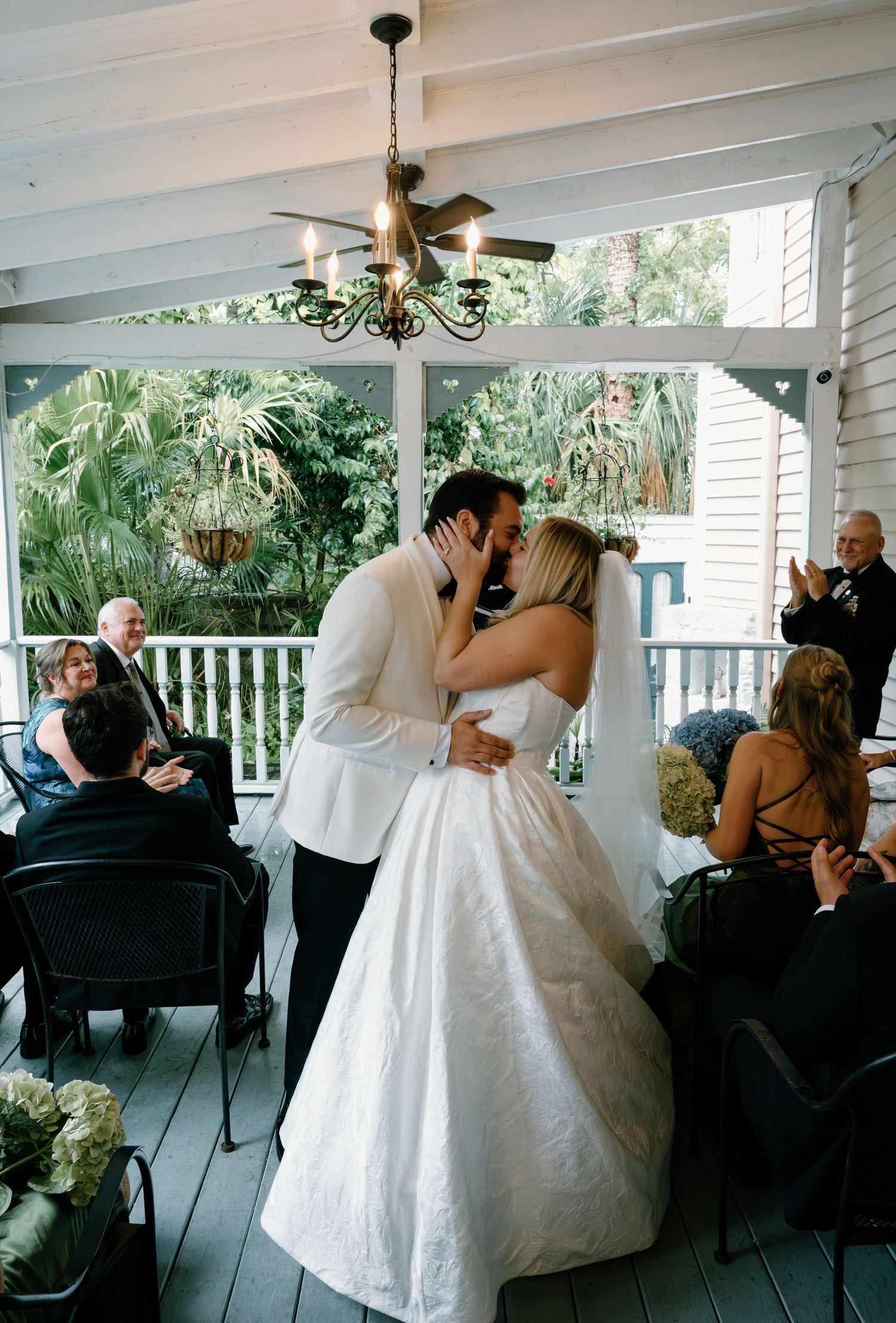 Bride and groom share first kiss during outdoor porch wedding ceremony with guests applauding.