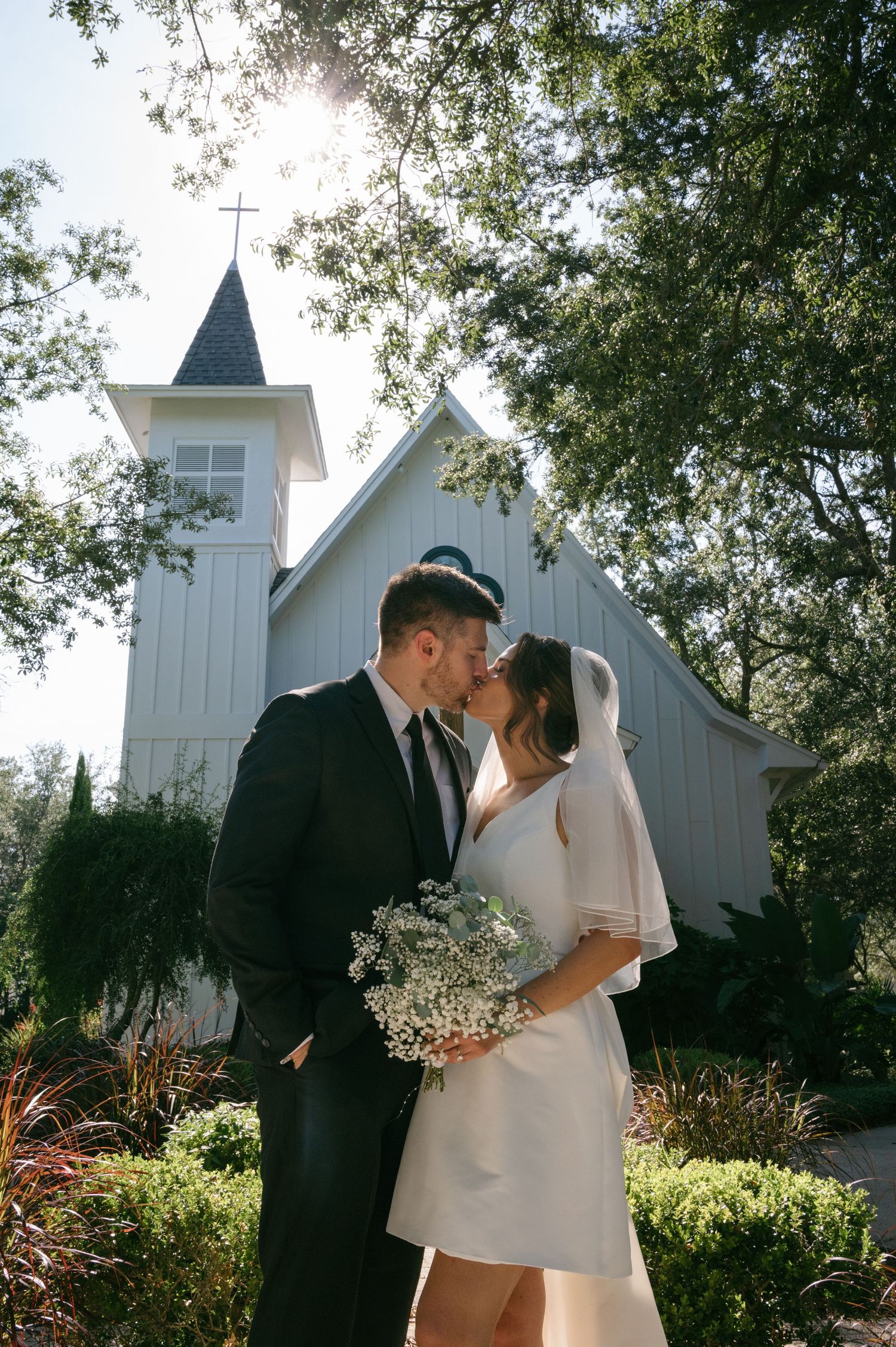Newlyweds kiss in front of a charming white chapel surrounded by trees, bride holding a baby's breath bouquet.