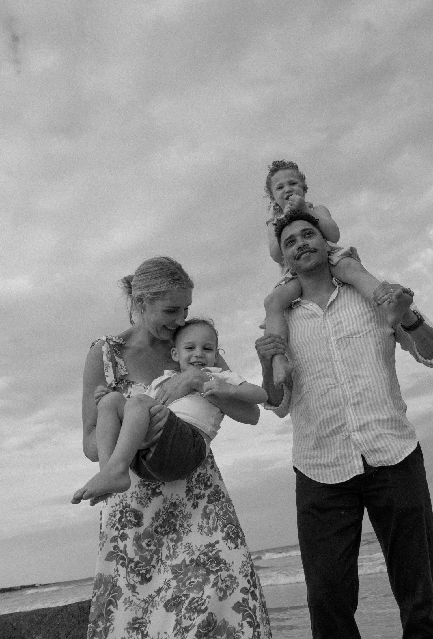 Black and white family portrait on beach, parents holding two young children with cloudy sky above.