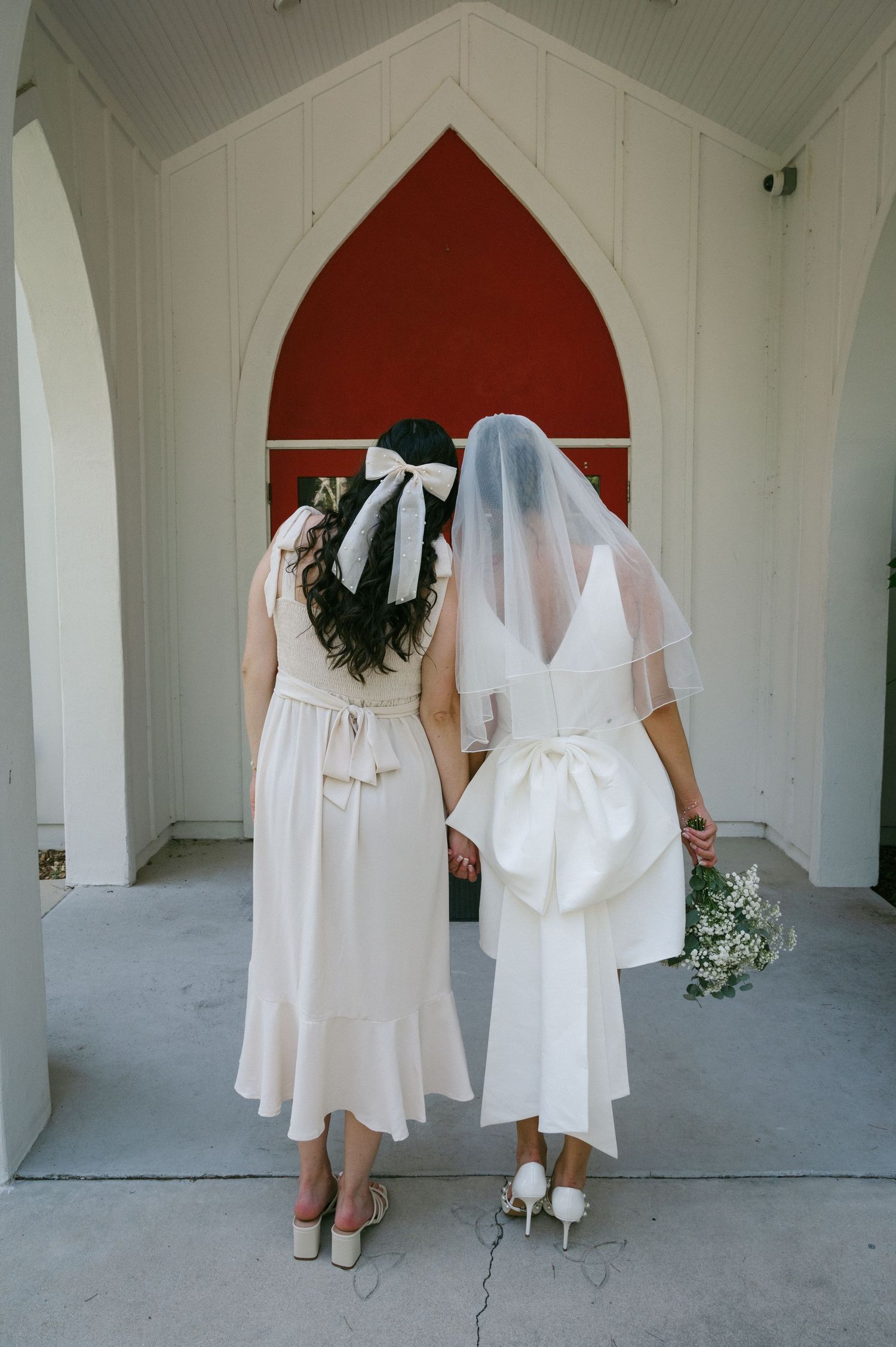 Bride and companion walking toward red arched church door, bride wearing veil and large bow on gown.