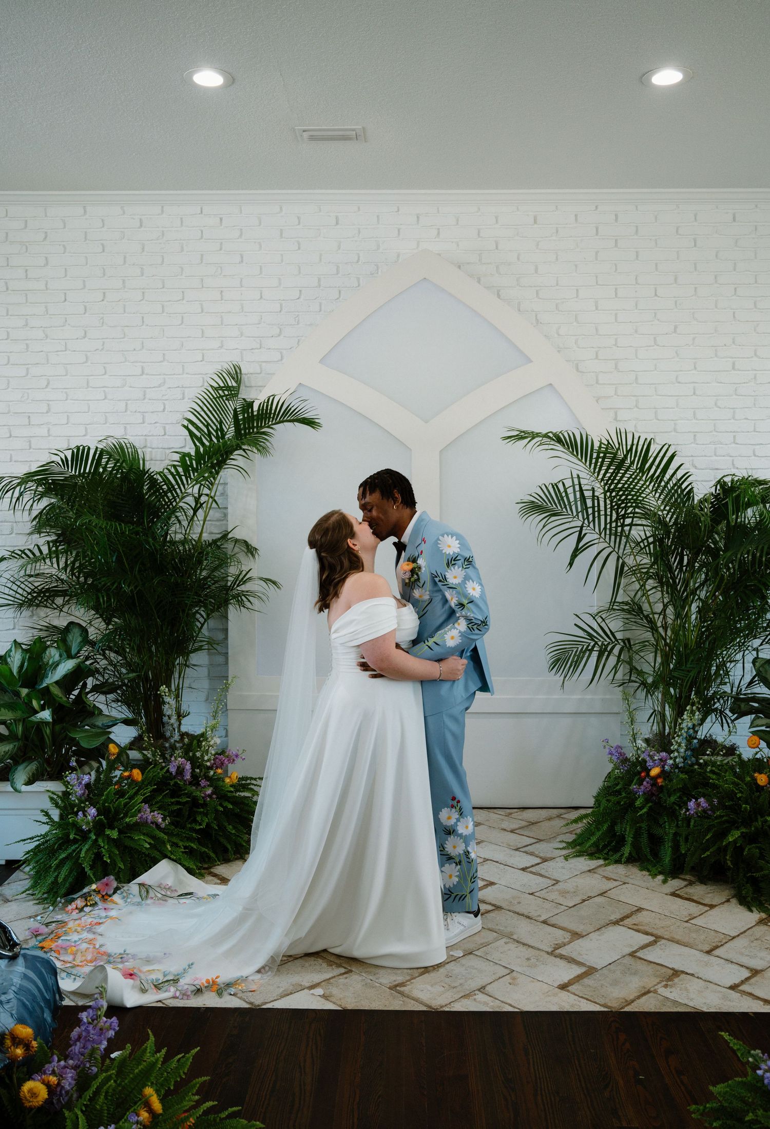 Bride and groom share first kiss at altar surrounded by tropical palms in white chapel.