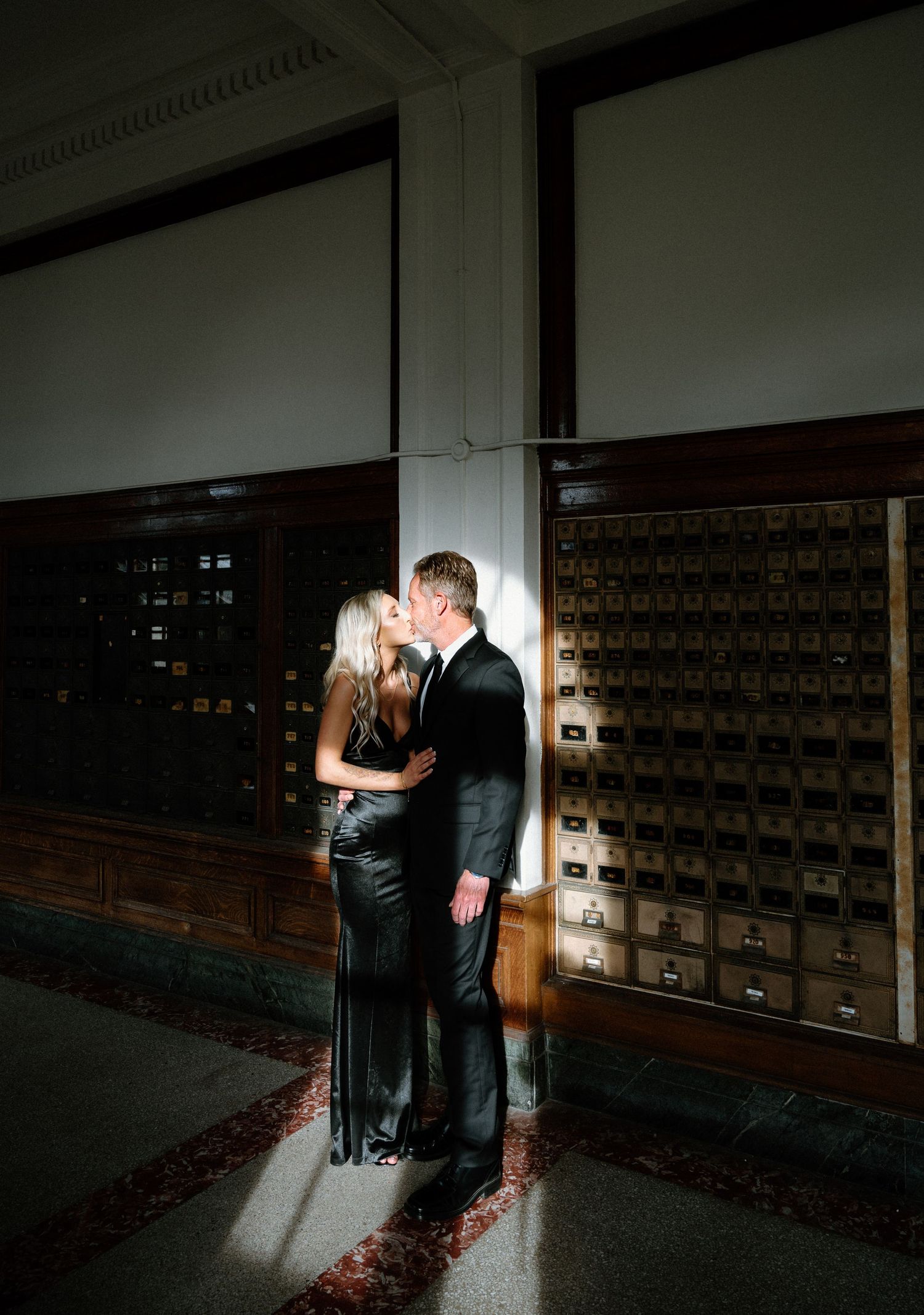 Elegant couple kissing in dramatic light beside vintage mailboxes in a formal indoor setting.