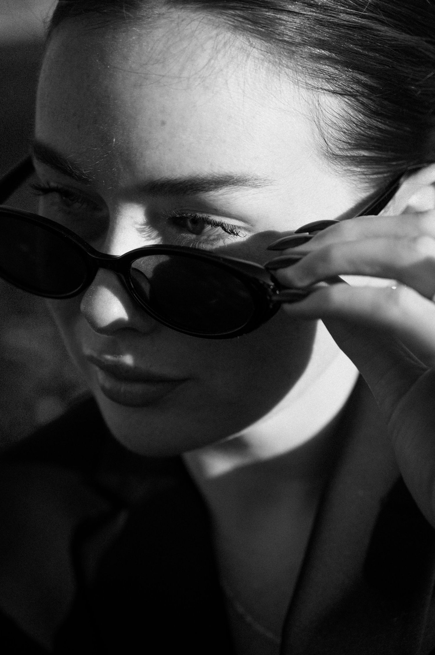 Close-up black and white portrait of woman adjusting oval sunglasses with dramatic shadow lighting.