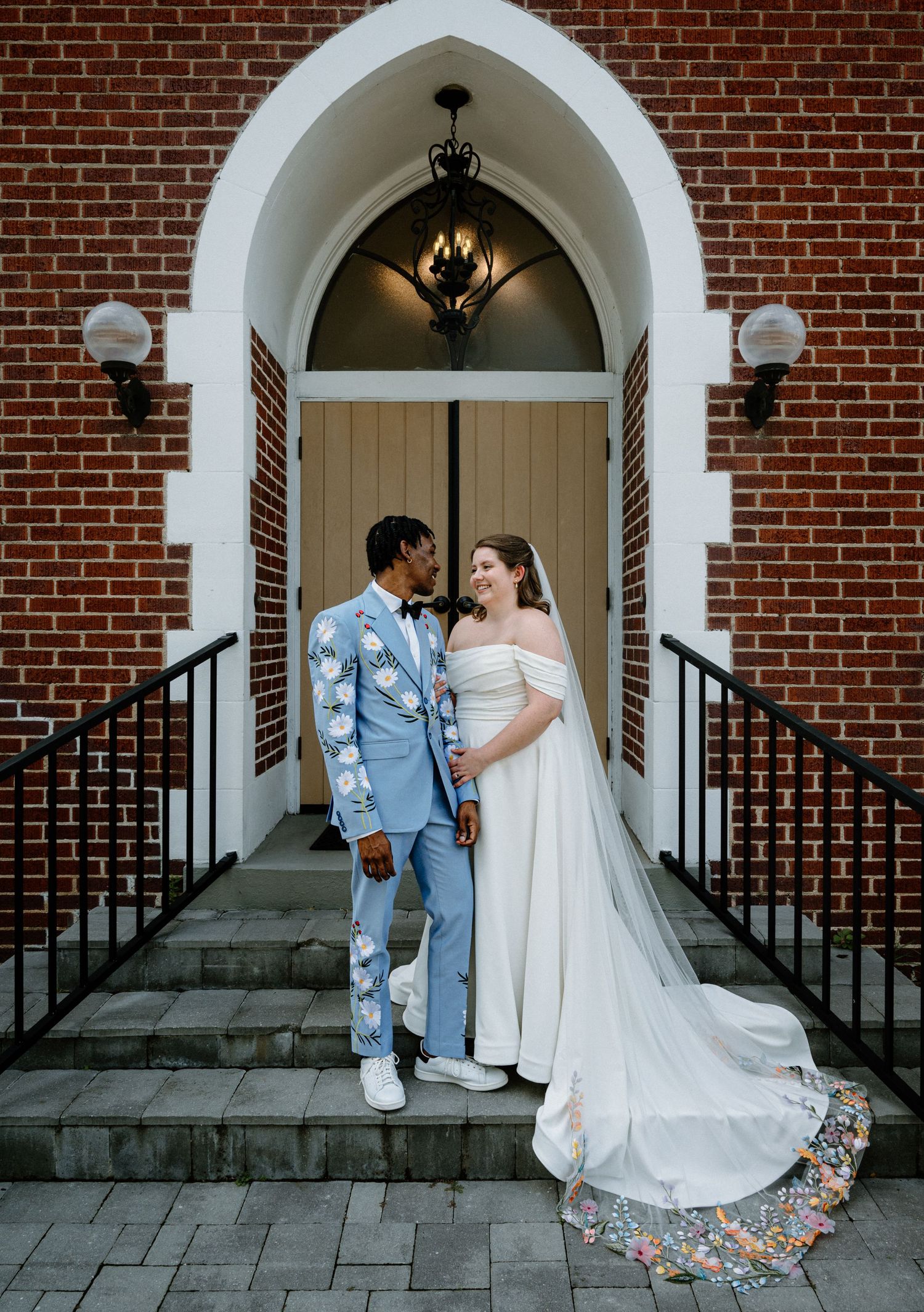 Couple in love poses on church steps, groom in blue floral suit, bride in white gown with floral cathedral train.
