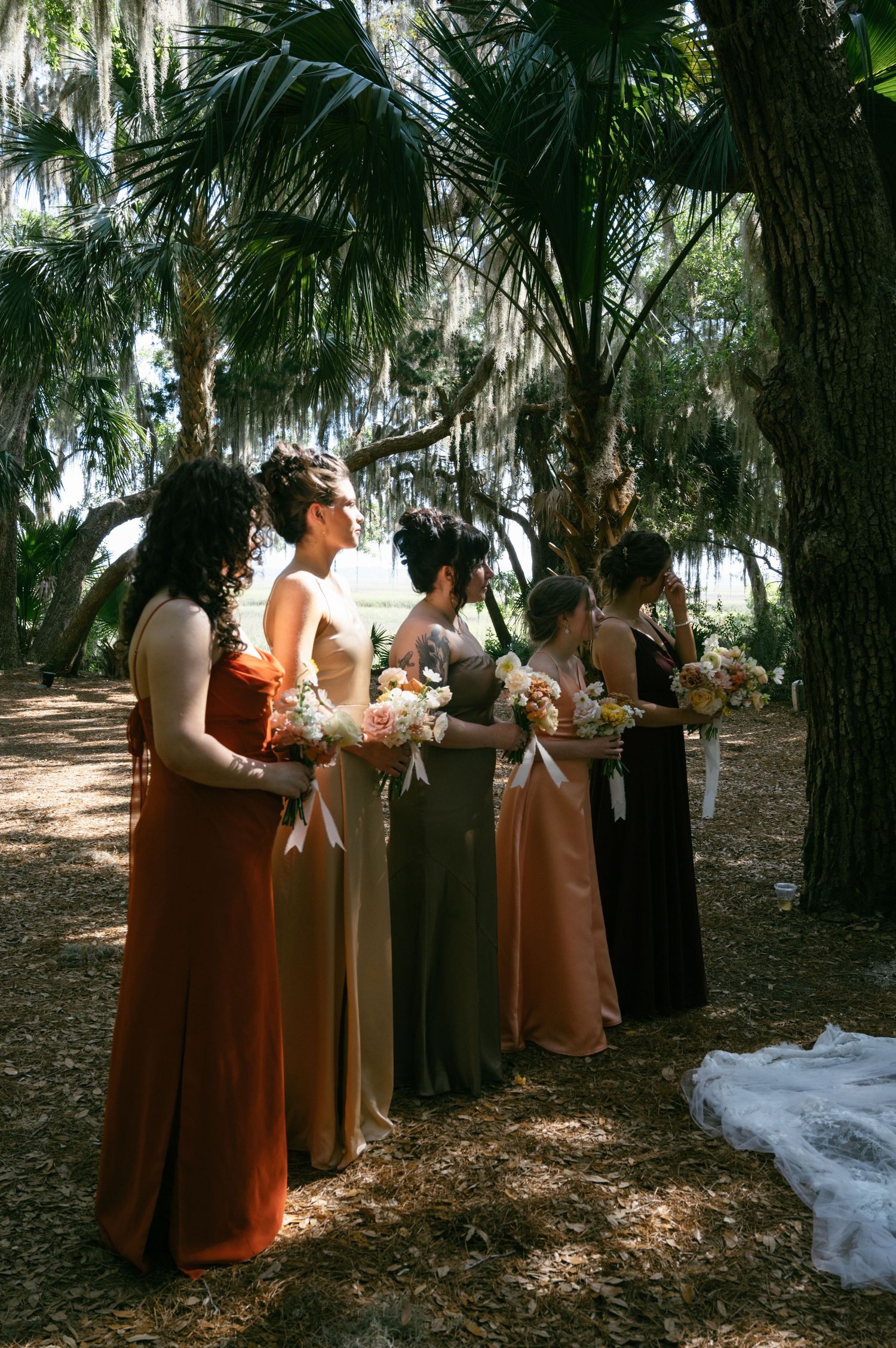 Bridesmaids in mismatched earth-tone gowns hold bouquets under Spanish moss-draped palms.