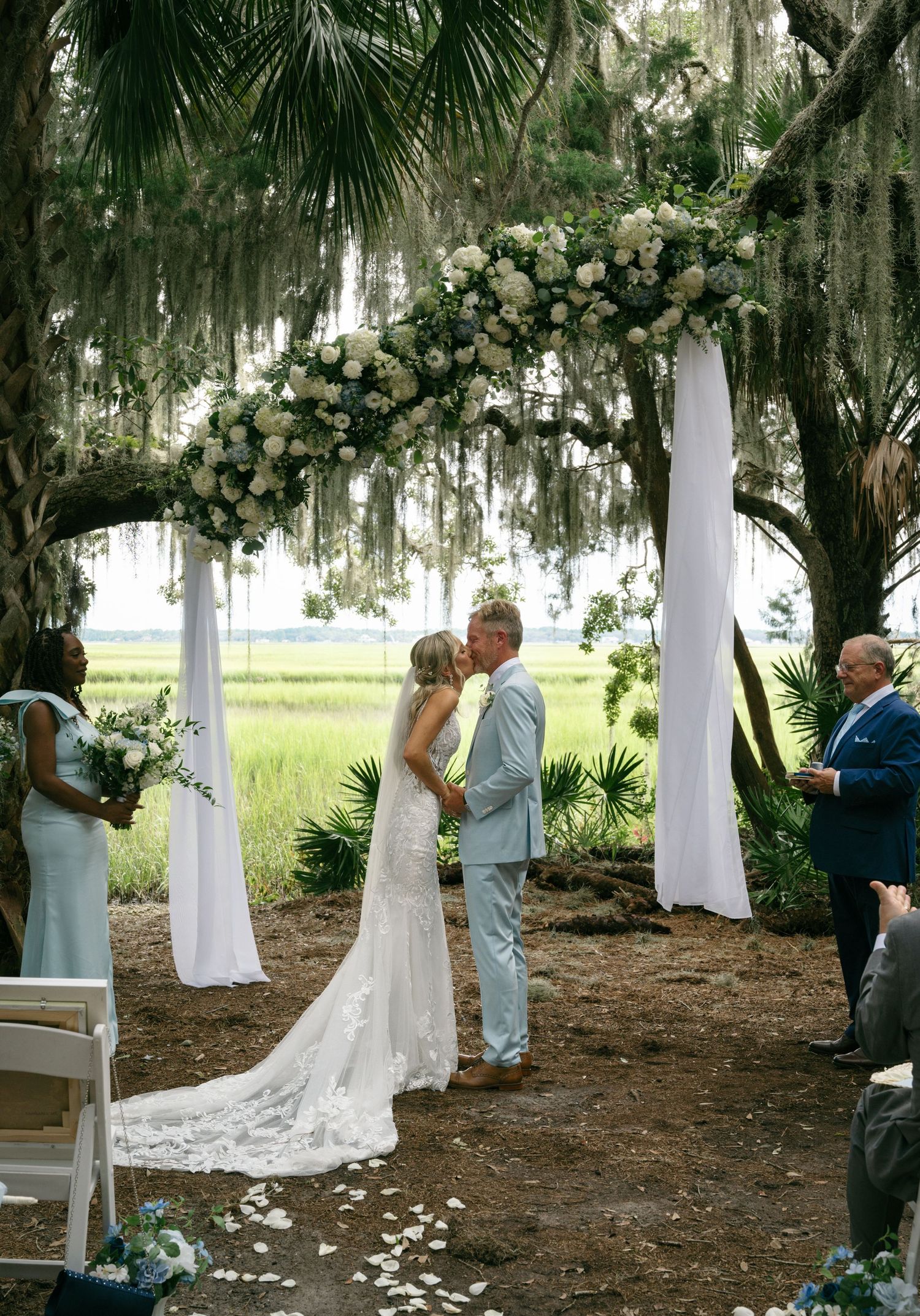 Bride and groom sharing a kiss under a floral arch draped with moss by a scenic marsh at Walker's Landing in Fernandina Beach, Florida.