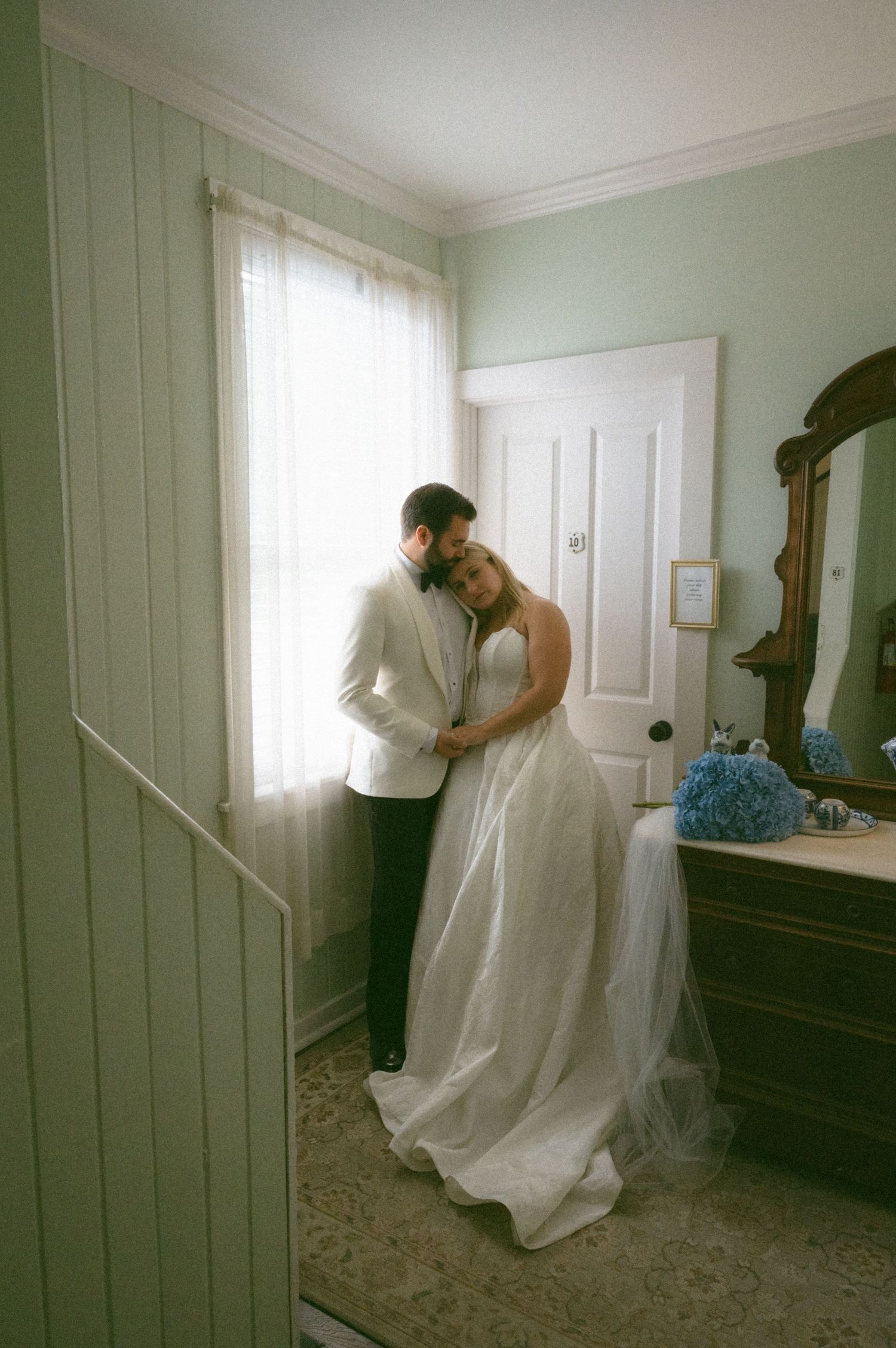 Bride and groom embracing tenderly by a window in a softly lit vintage bedroom.