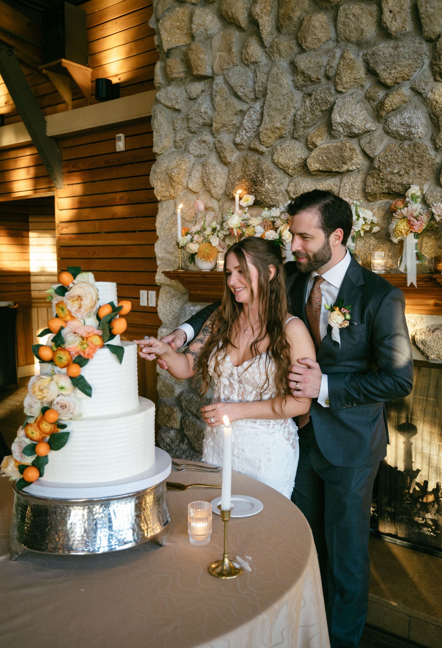 Smiling bride and groom cut white tiered wedding cake decorated with oranges and flowers.