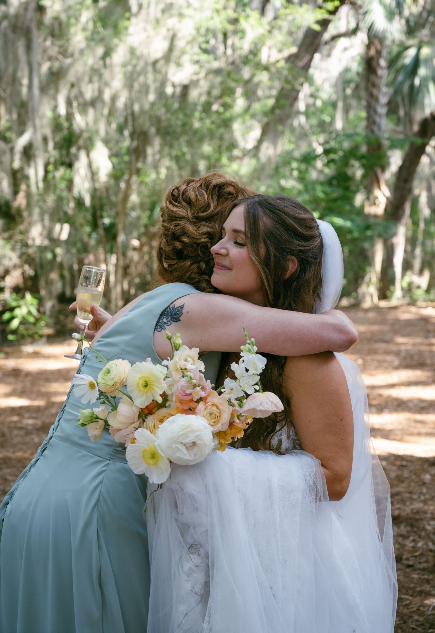 Bride embraces bridesmaid holding bouquet and champagne under moss-covered trees.