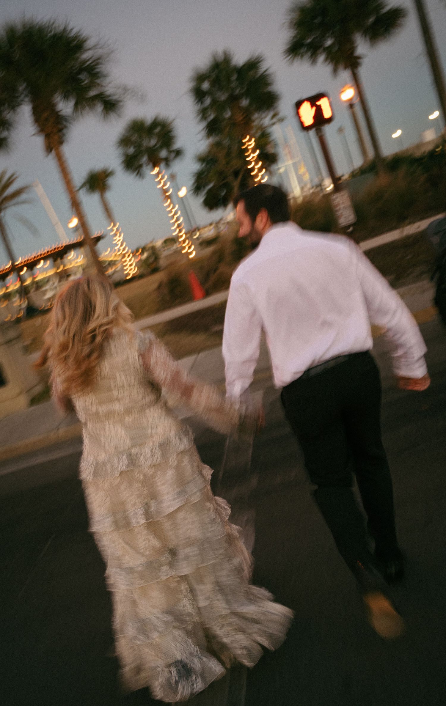 Blurred motion shot of bride and groom running hand in hand past palm trees at dusk.