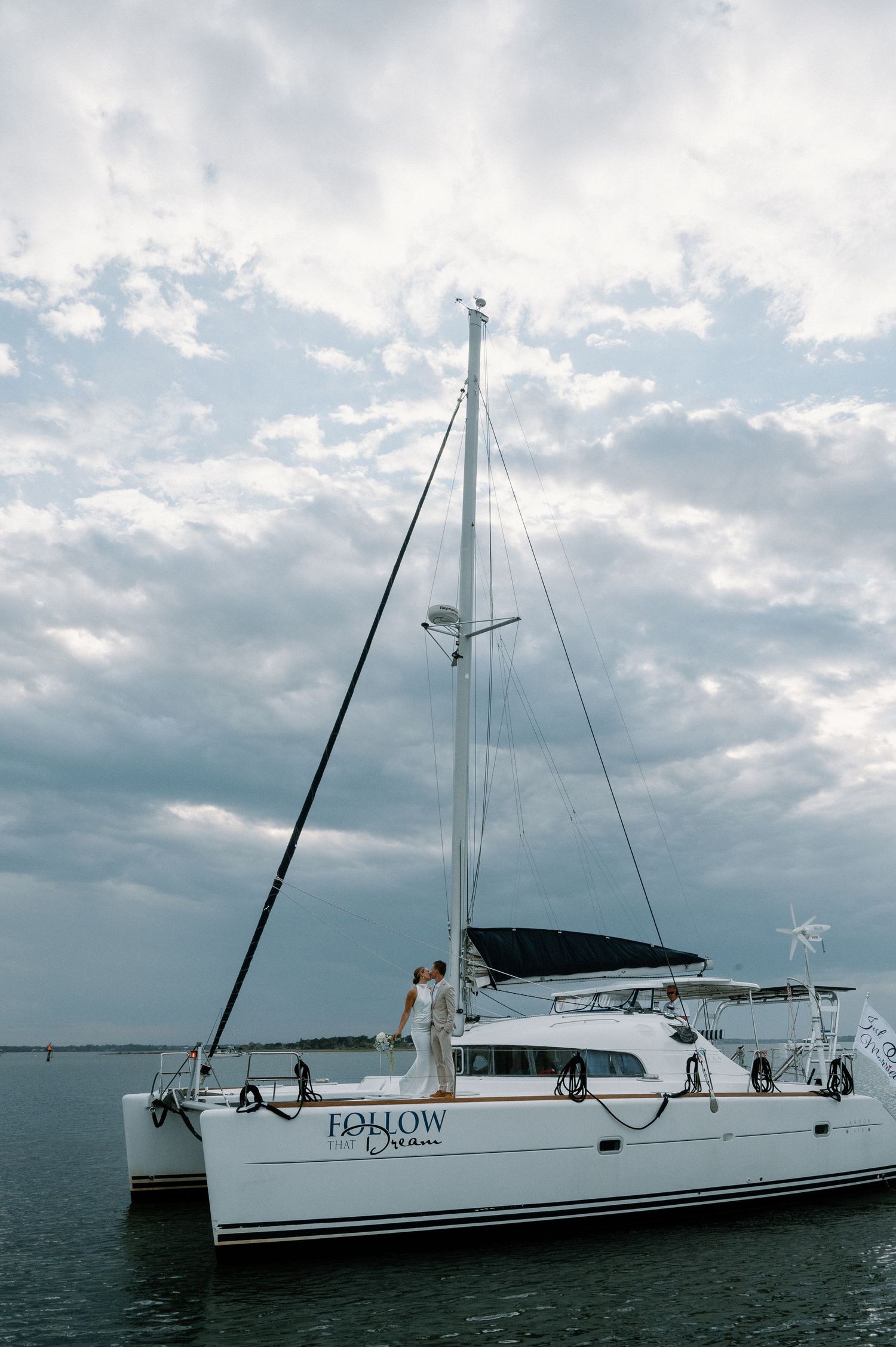 Wedding couple standing on a white catamaran named Follow Dream under dramatic cloudy skies.