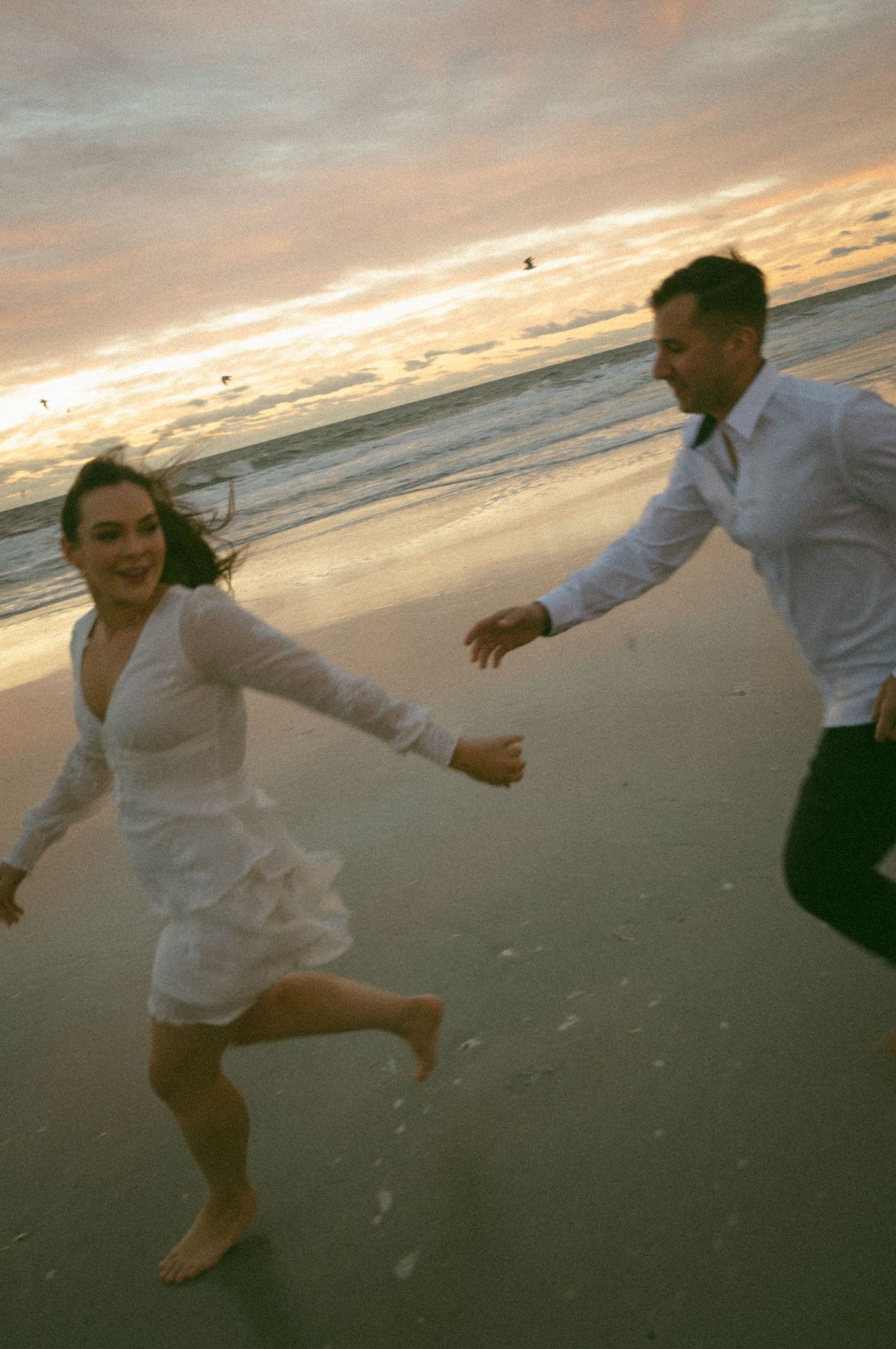 Couple in white running joyfully on a beach at sunset, waves and colorful sky in the background.