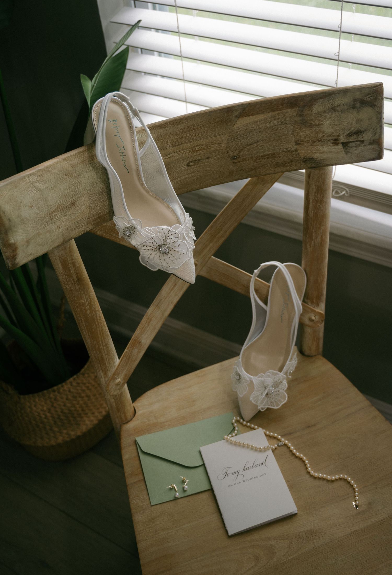 White bridal heels with floral detail displayed on wooden chair beside pearl necklace and green envelope.