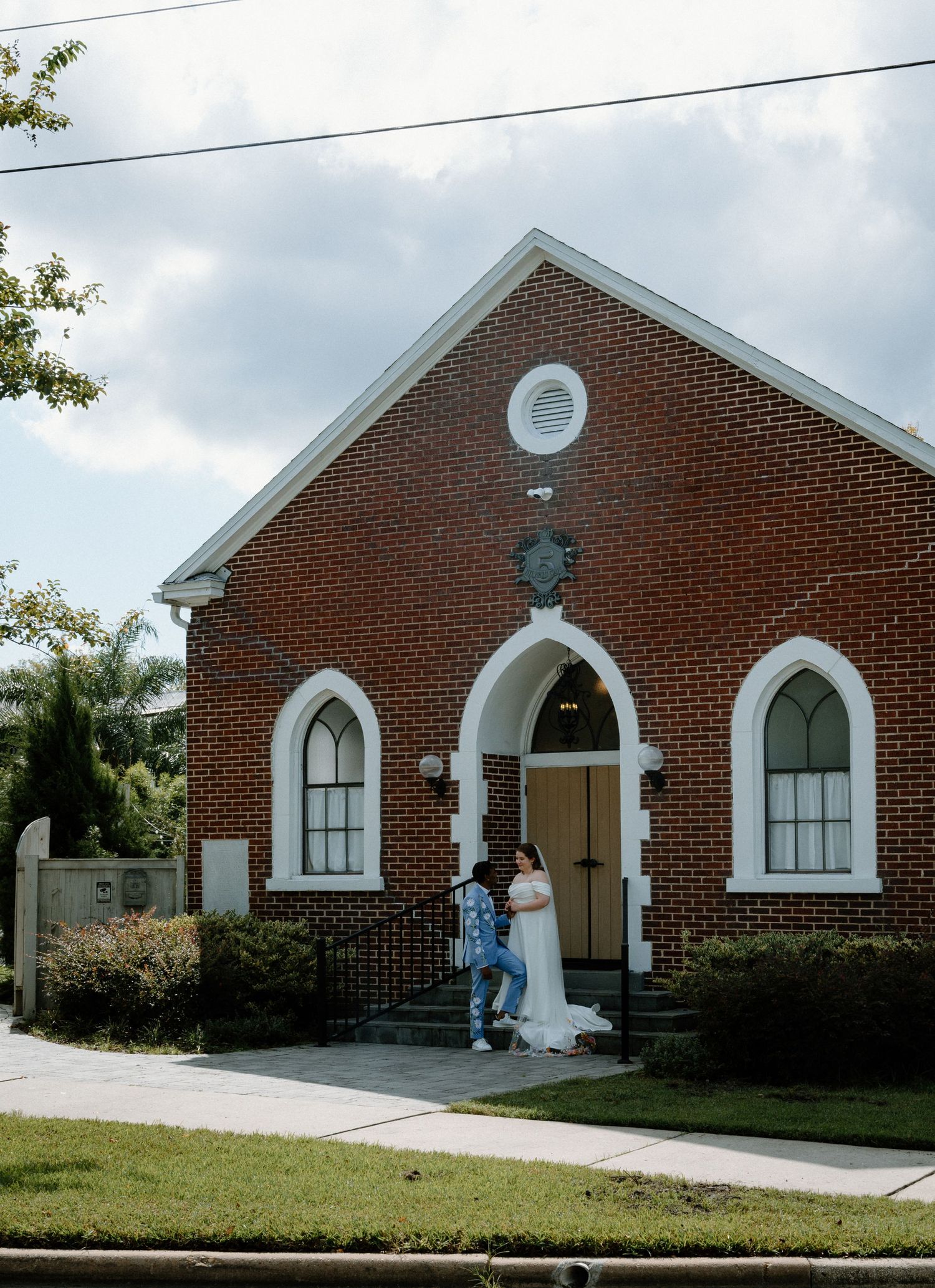Bride and groom pose outside charming red brick chapel with white arched windows and wooden door.