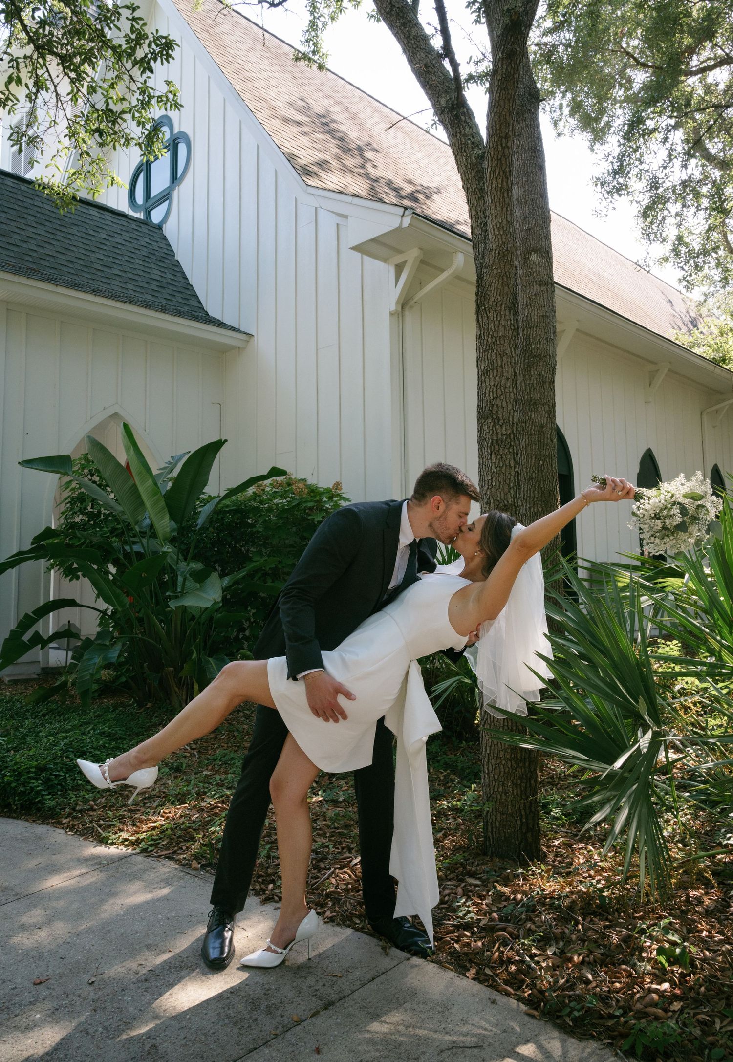Groom dipping bride in white mini dress and veil outside white chapel surrounded by tropical greenery.