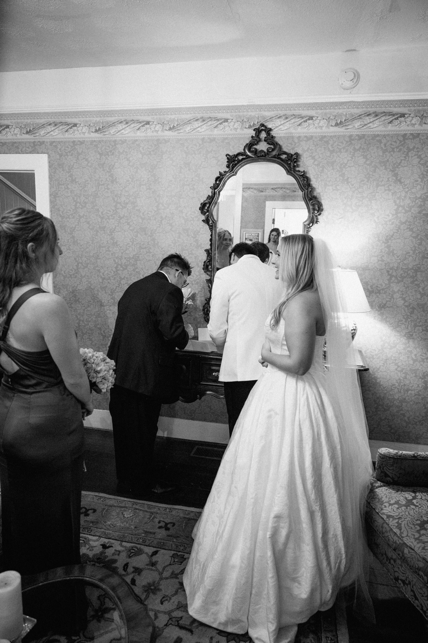 Black and white photo of bride and groom signing marriage documents before ornate mirror.
