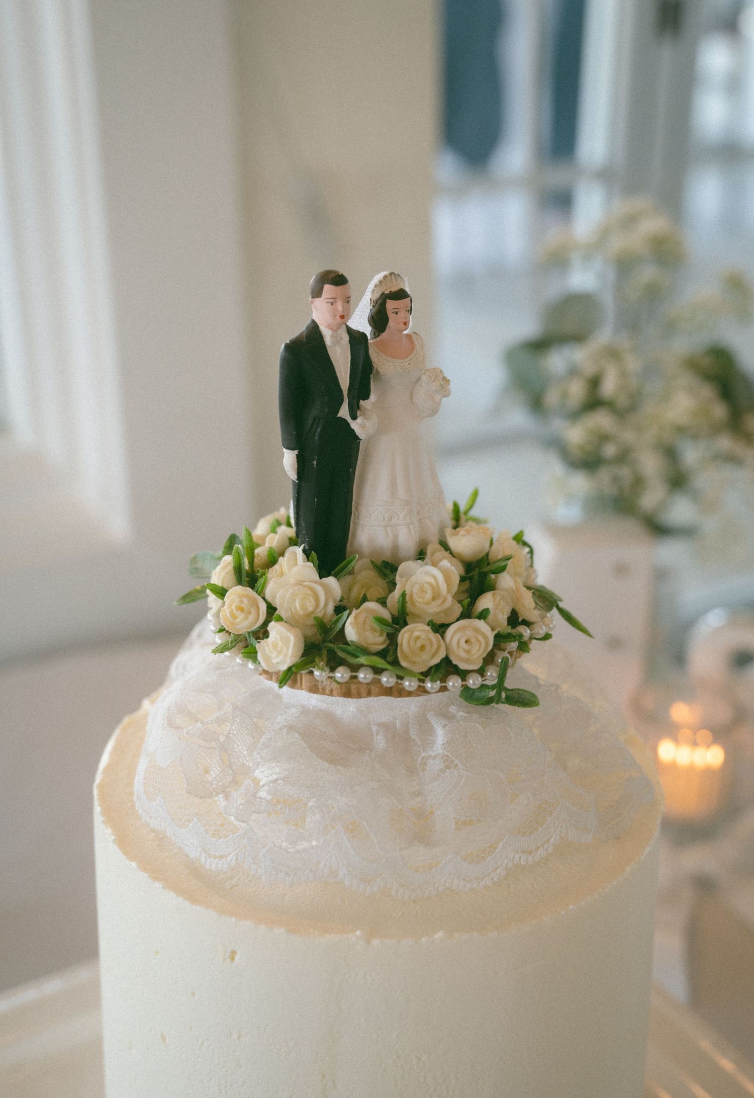 Elegant wedding cake topped with classic bride and groom figurines surrounded by white roses and lace trim.