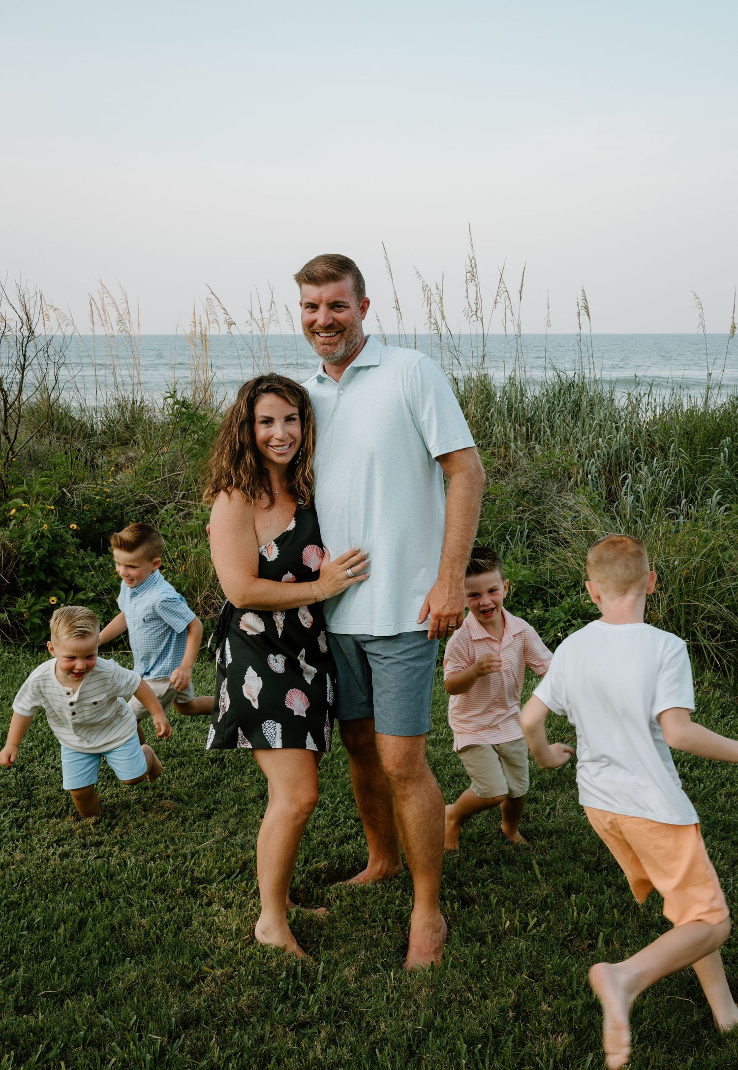 Happy family with three energetic boys posing together near a scenic ocean beach at dusk.