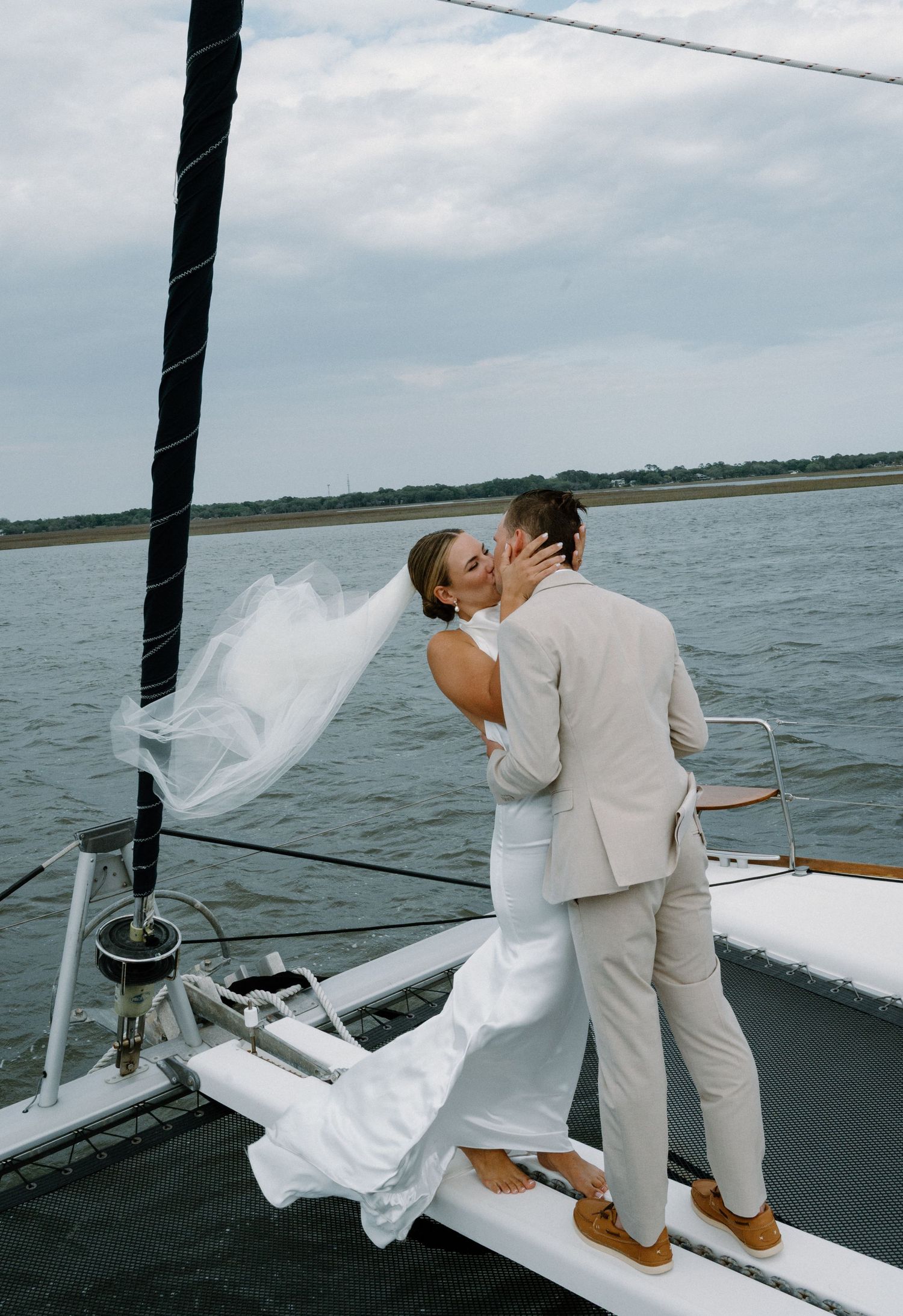 Bride and groom sharing a kiss on sailboat bow with veil flowing in the coastal breeze.