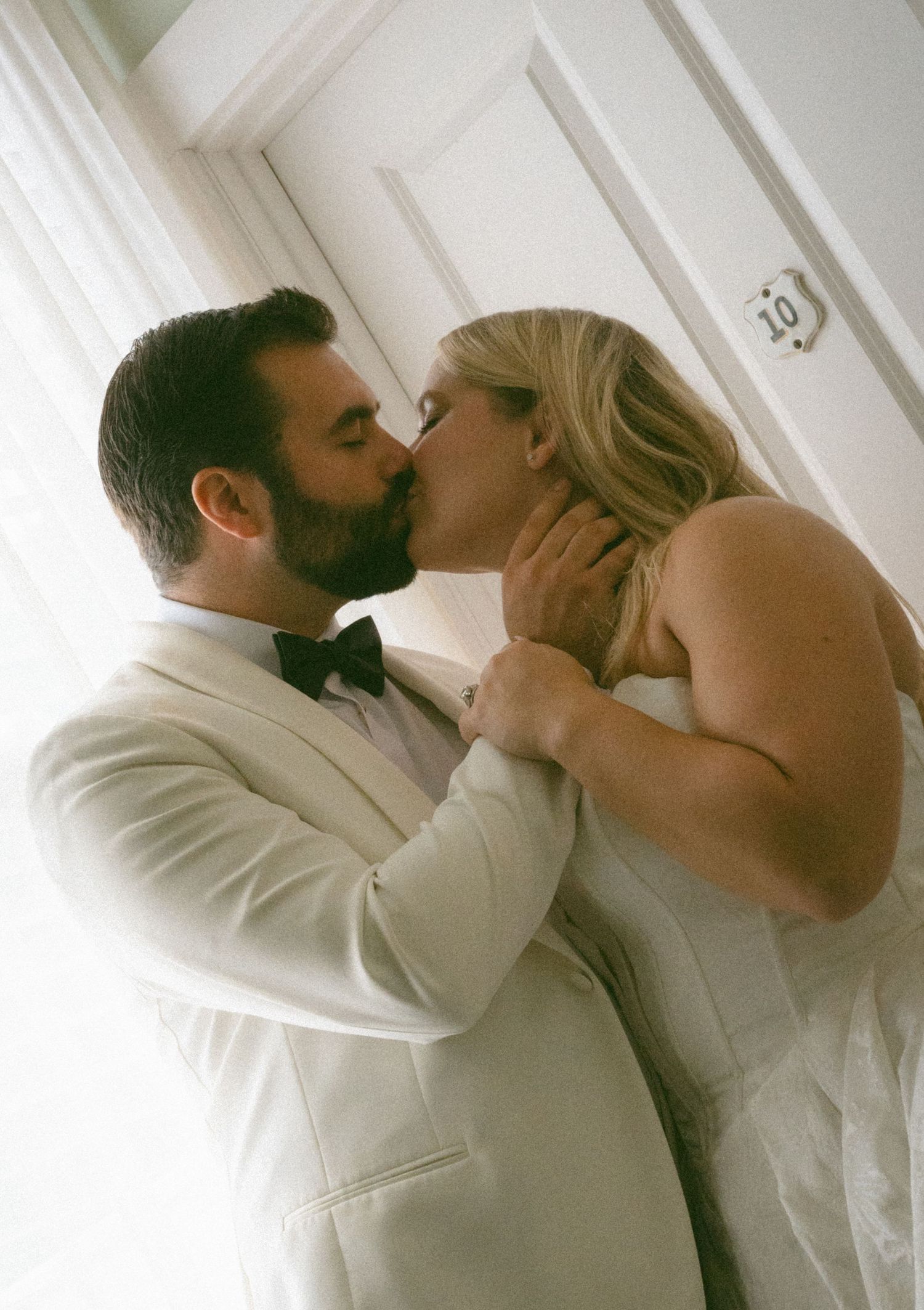 Bride and groom sharing a romantic kiss in front of a white door in soft light.