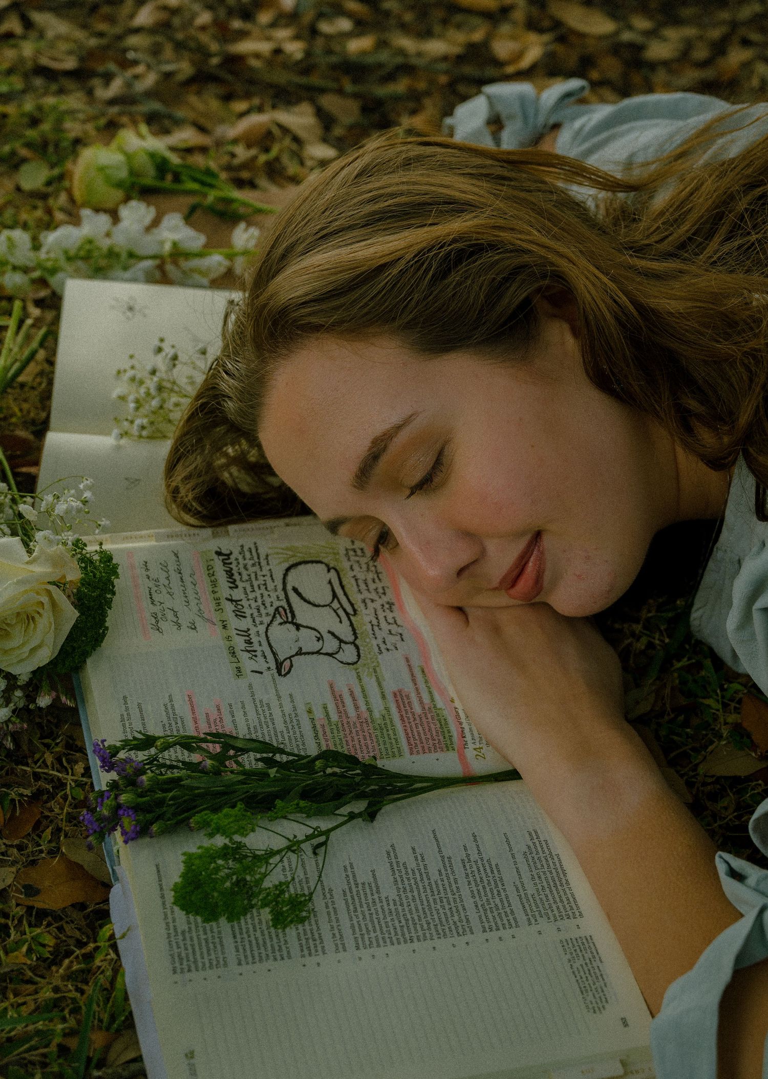 Young woman resting her head on an open annotated Bible surrounded by wildflowers outdoors.