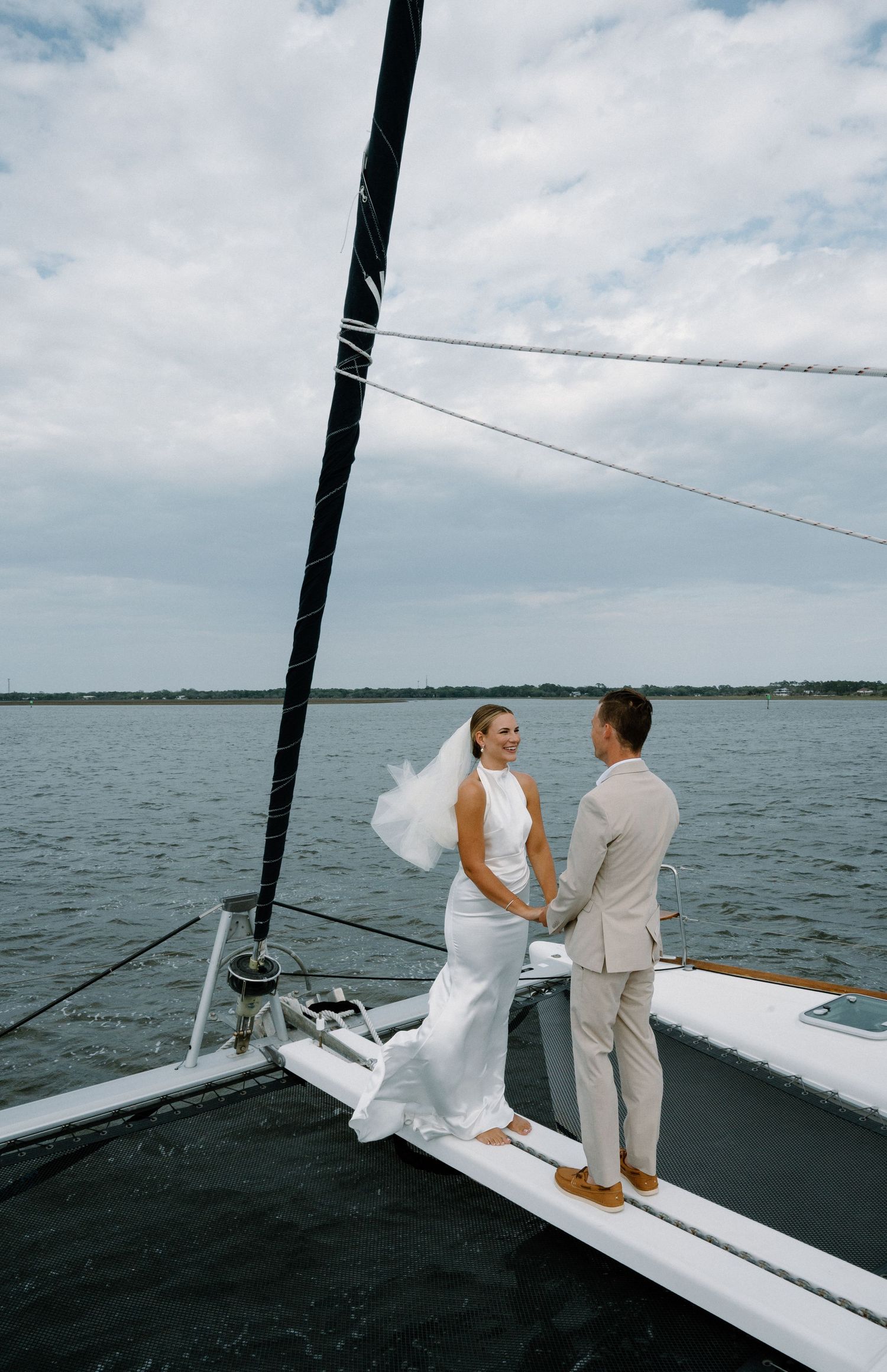 Bride and groom holding hands on catamaran bow with cloudy sky and open water behind them.