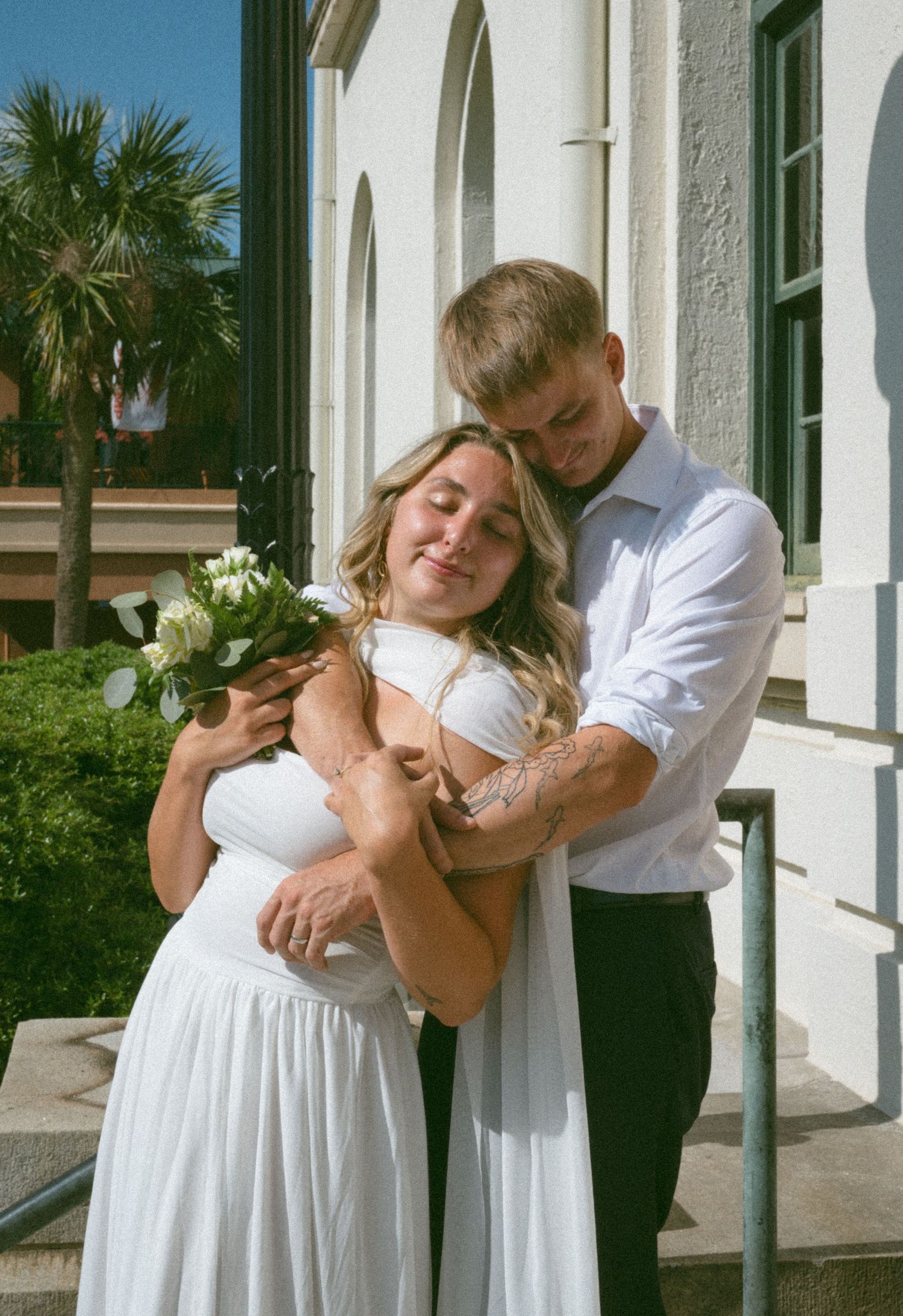 Groom tenderly embraces bride holding white bouquet on sunlit steps of elegant white building.