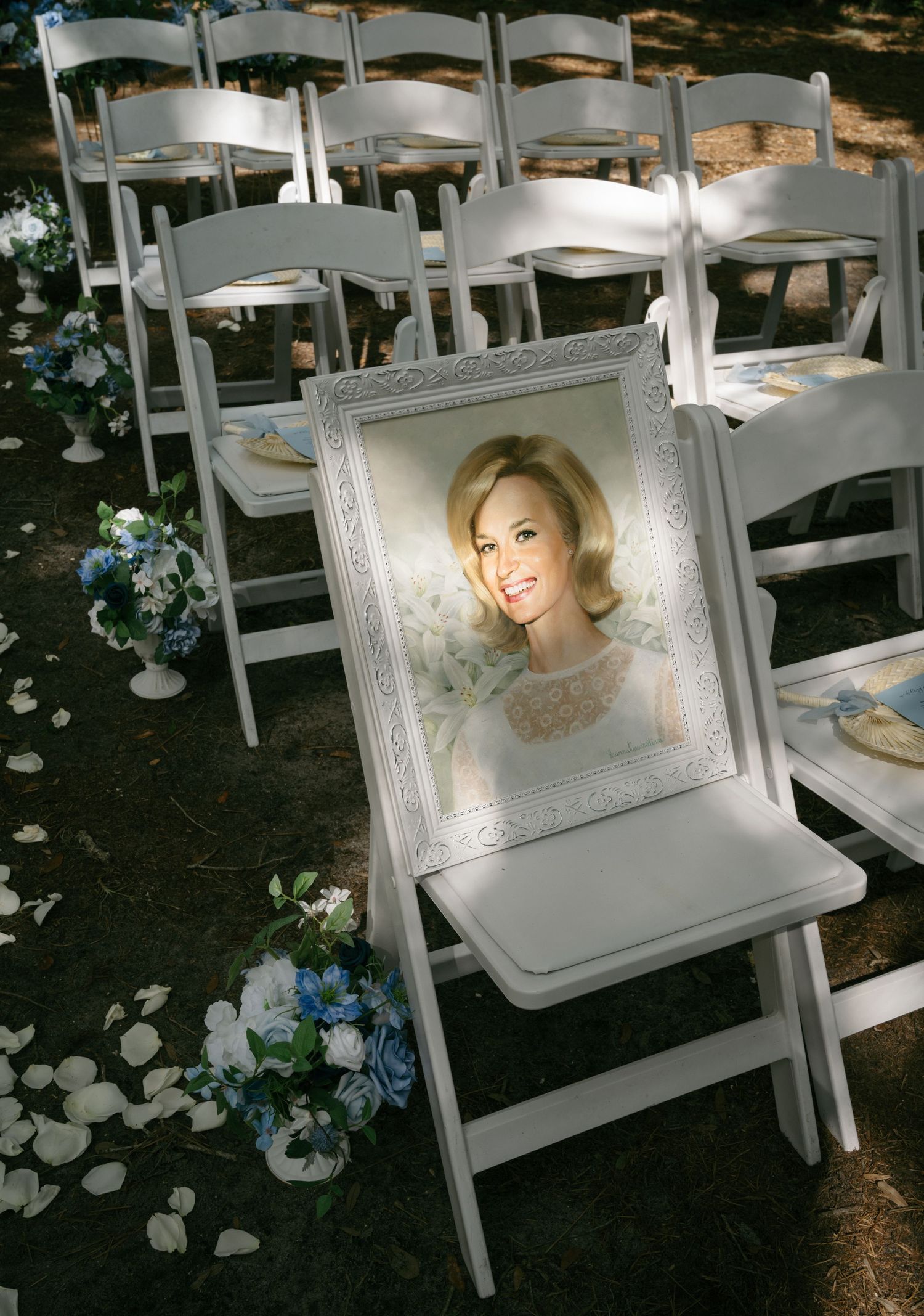 Framed memorial photo of loved one placed on white chair at outdoor wedding ceremony aisle.