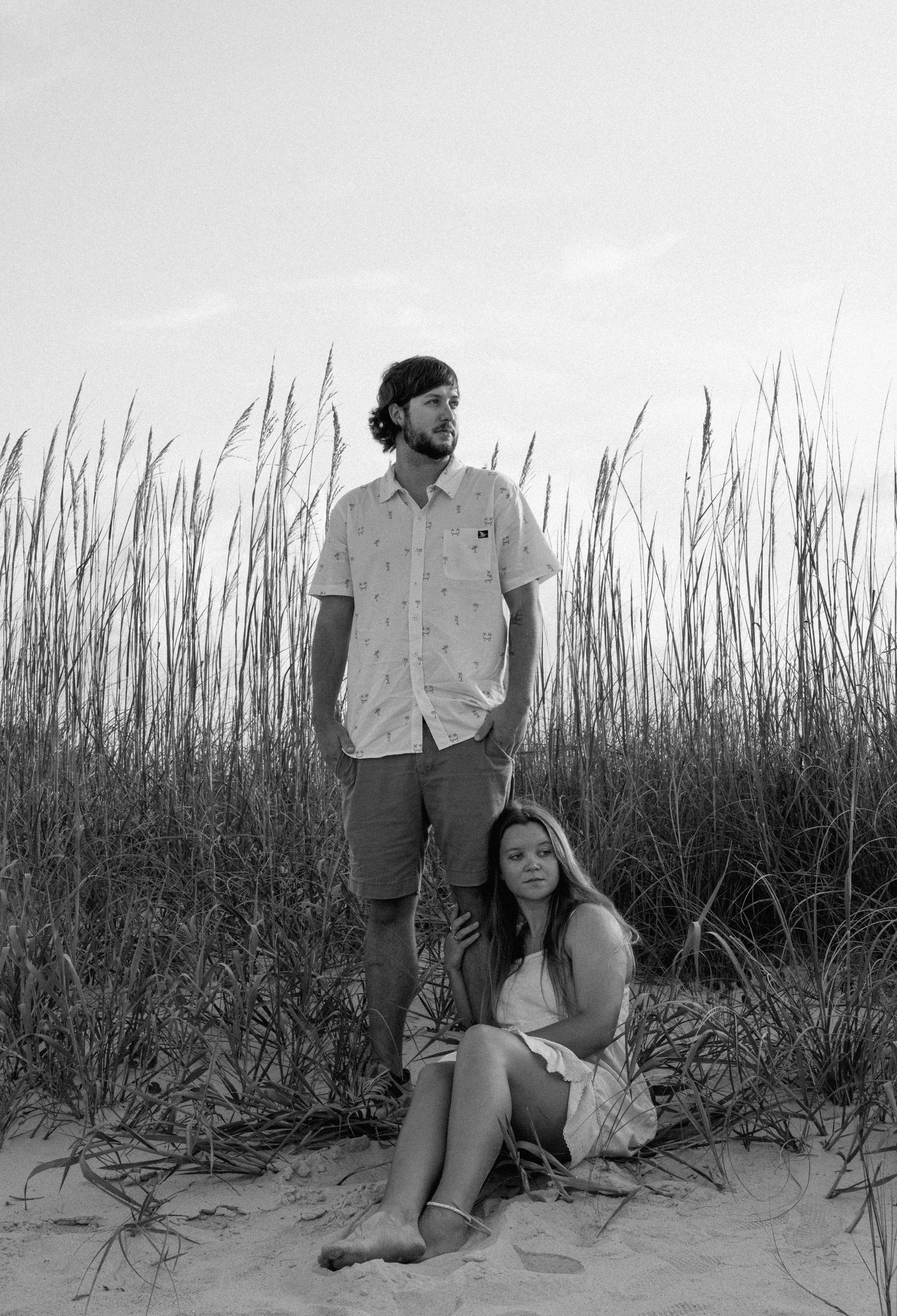 Black and white photo of a couple posing on sandy beach dunes surrounded by tall grass.